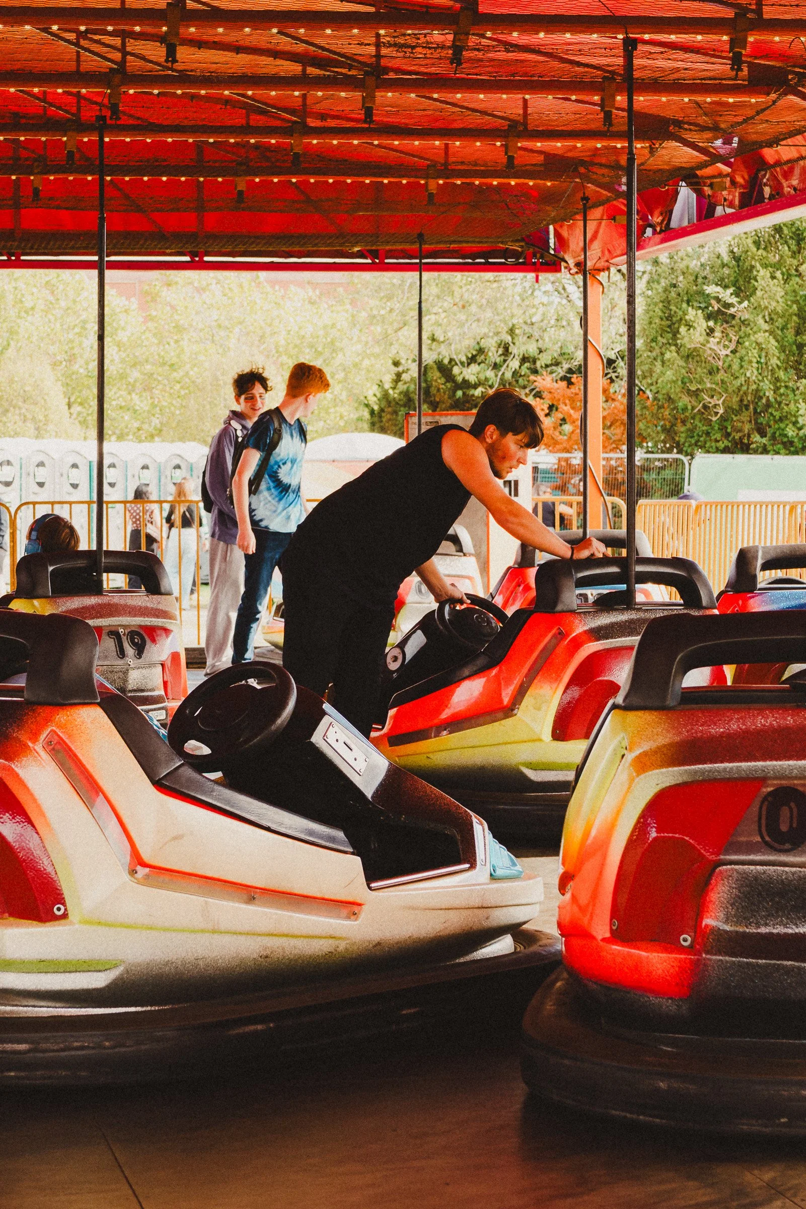 Two young men and a young woman at an amusement park ride, with the men sitting in bumper cars and the woman standing next to them, under a wooden canopy with string lights and trees visible in the background.