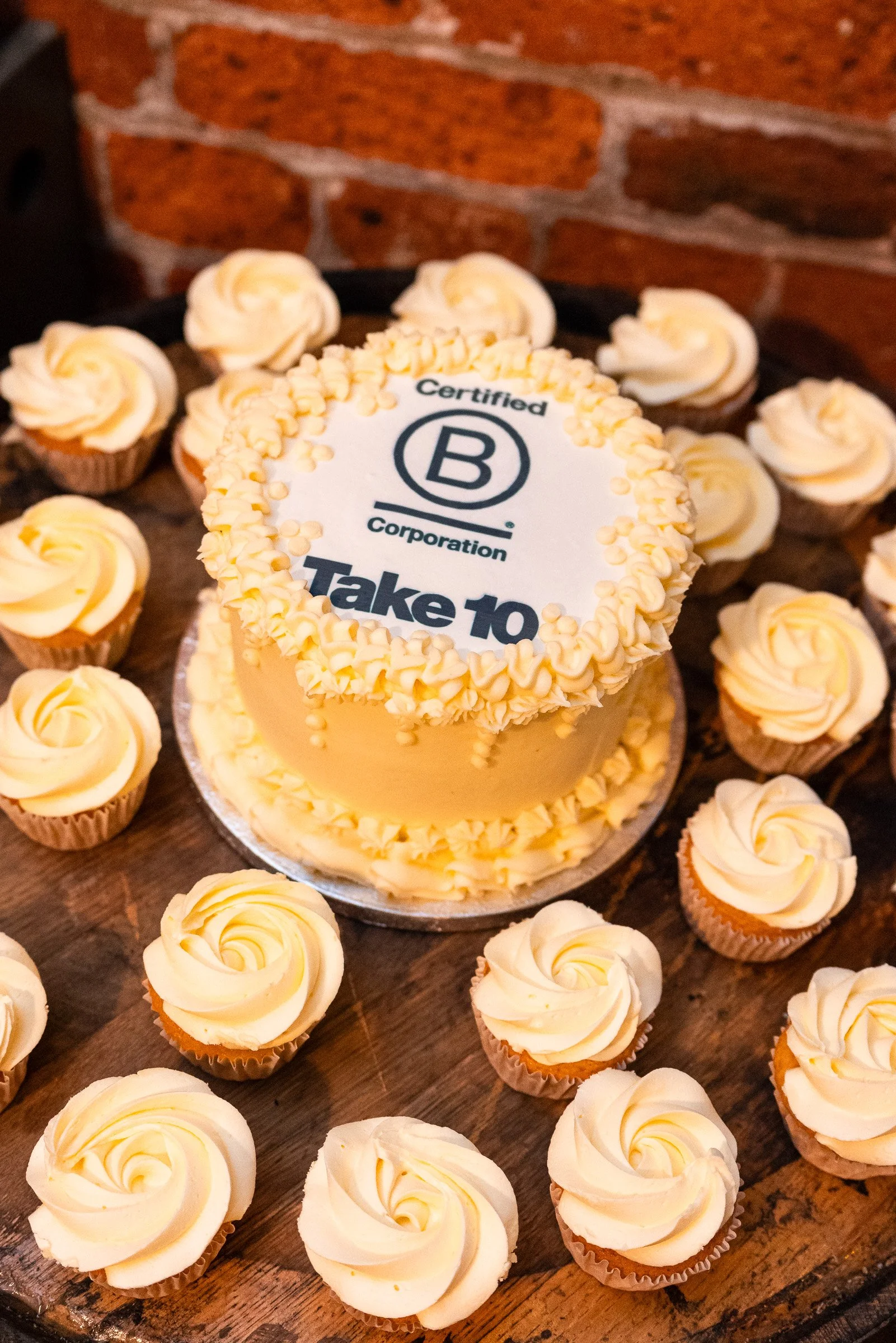 A cake with a white frosting and the logo of Certified B Corporation with the text "Take 10" on top, surrounded by cupcakes with white swirl frosting on a wooden surface.