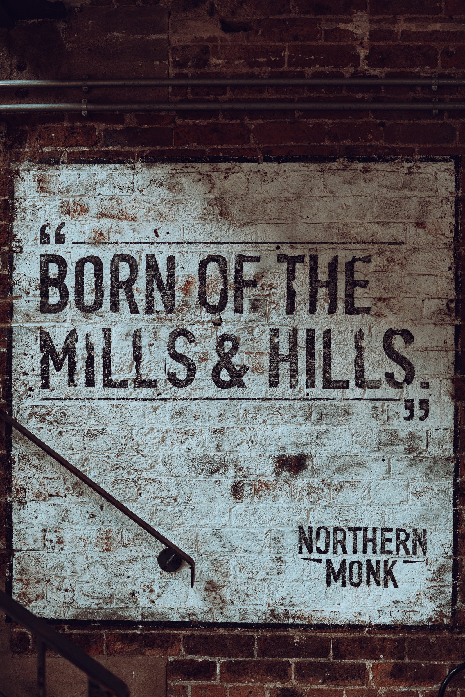 A white brick wall with black text: "BORN OF THE MILLS & HILLS." and a smaller text below reading "NORTHERN MONK." The wall is part of an industrial setting with exposed red brick and electrical conduits.