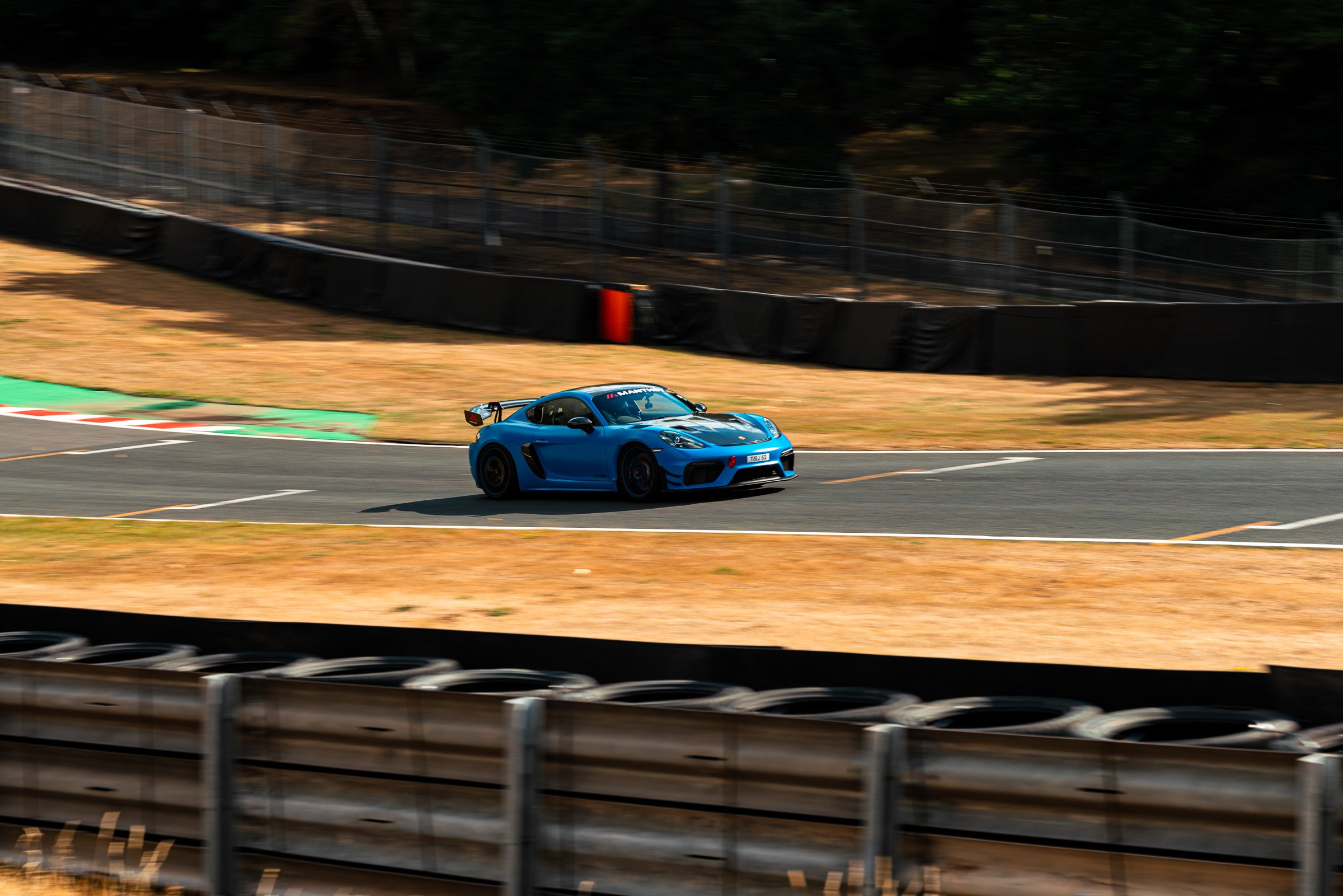 A blue Porsche race car on a racetrack during daylight, with tire barriers and fencing in the background.