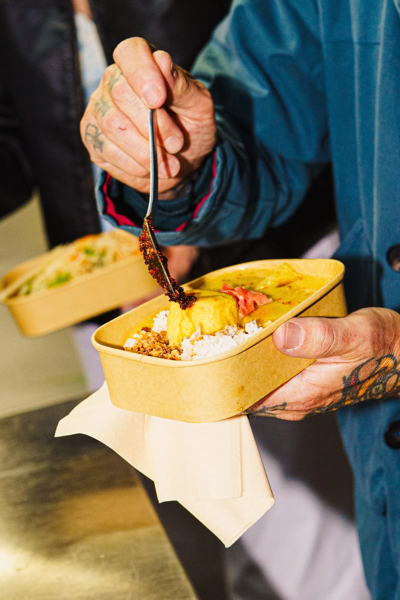 Person holding a takeout container of food with rice, an omelet, and meat. Someone else in the background is also holding a container of food.