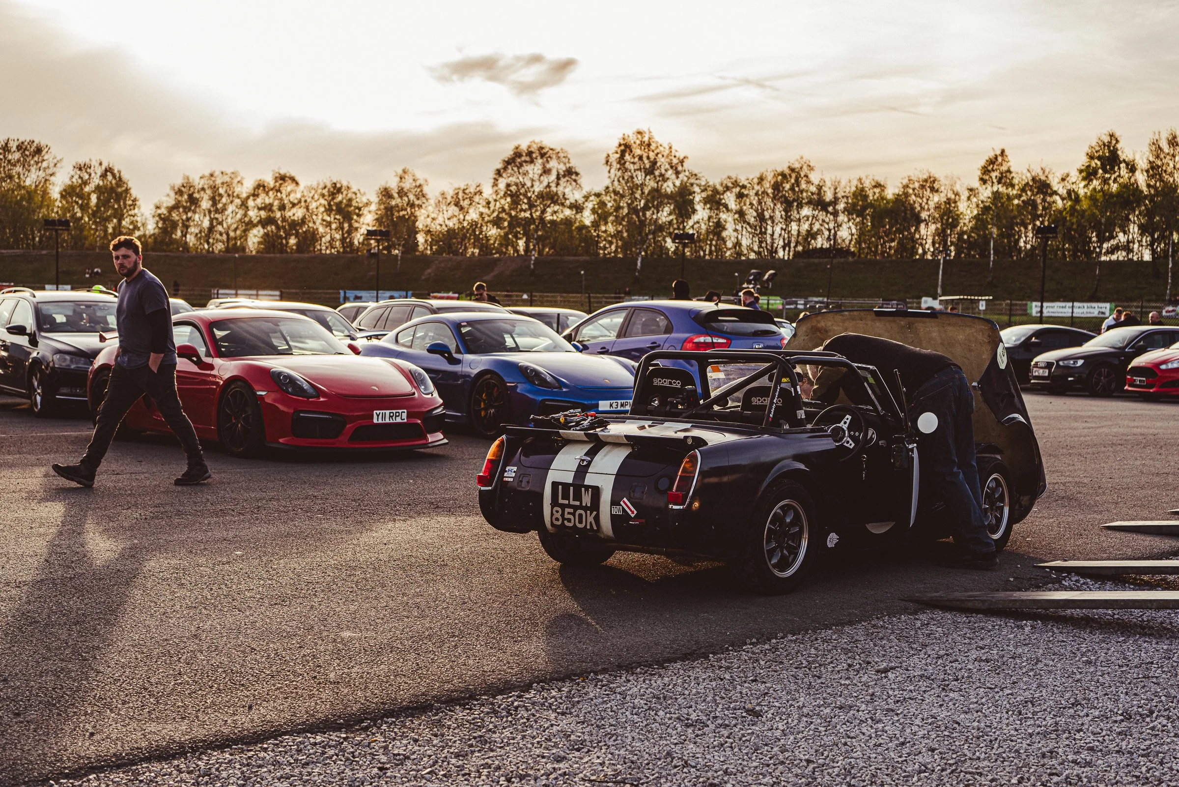 A parking lot with various sports cars, including a red Porsche, blue Porsche, and black convertible, during sunset with a few people around, and trees in the background.