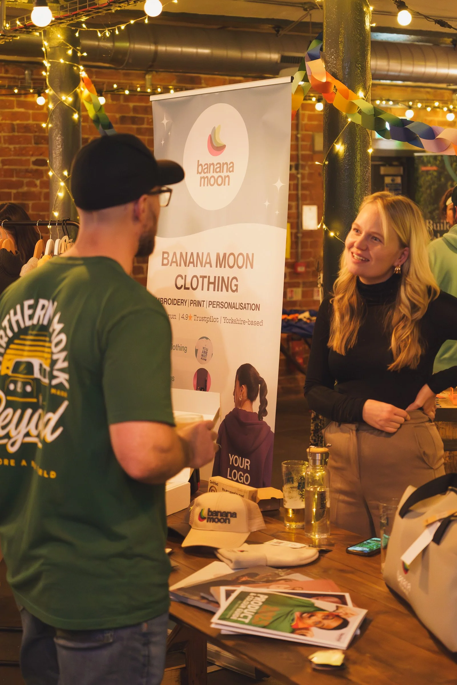 Two people talking at a promotional booth for Banana Moon clothing, with a banner behind them and a table displaying magazines, brochures, and merchandise, in a warmly lit indoor setting decorated with string lights and paper chain garlands.