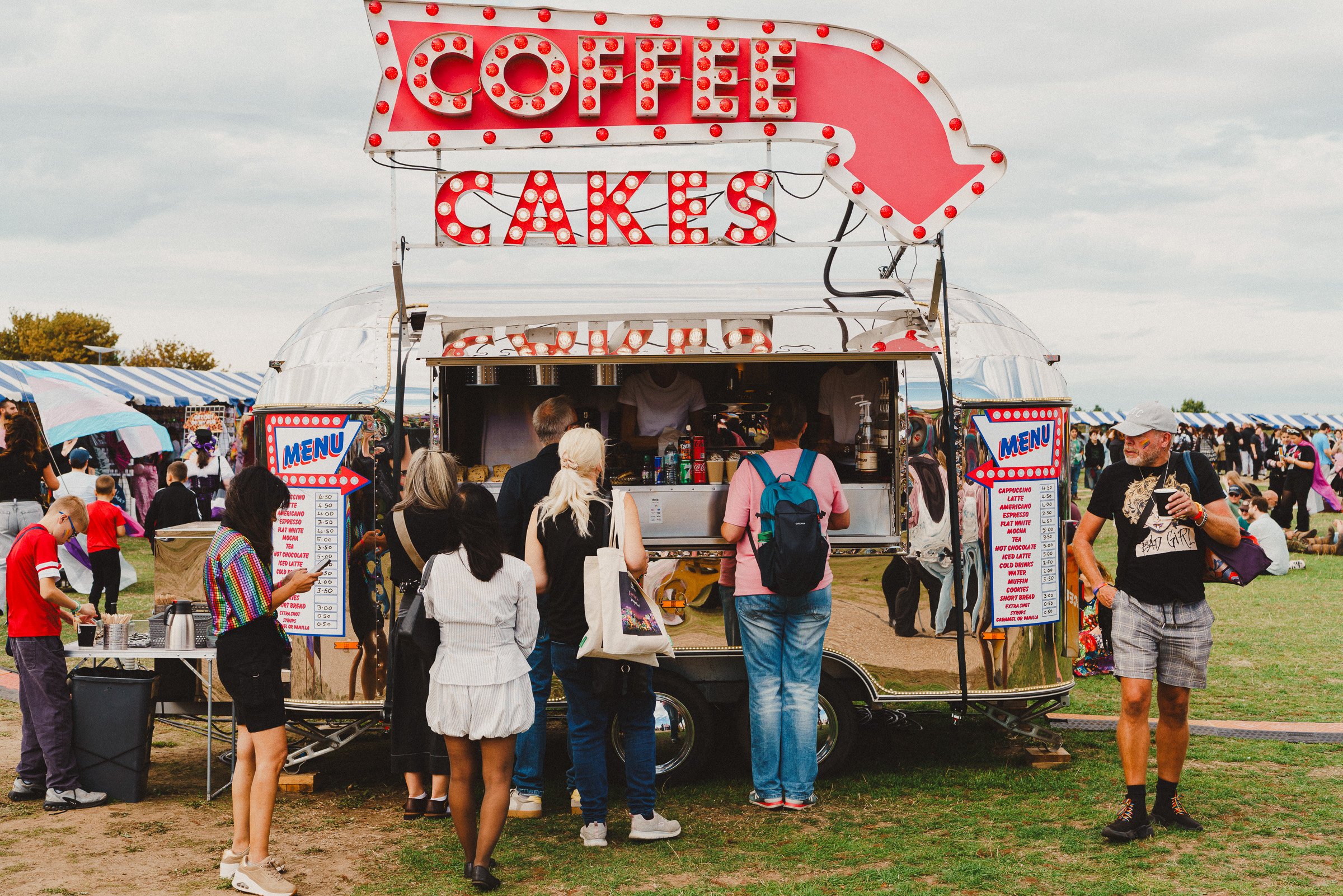 A crowded outdoor fair with people gathered in front of a coffee and cakes stand. People are ordering drinks and snacks from the stand, and others are standing in line or walking around the fairground.