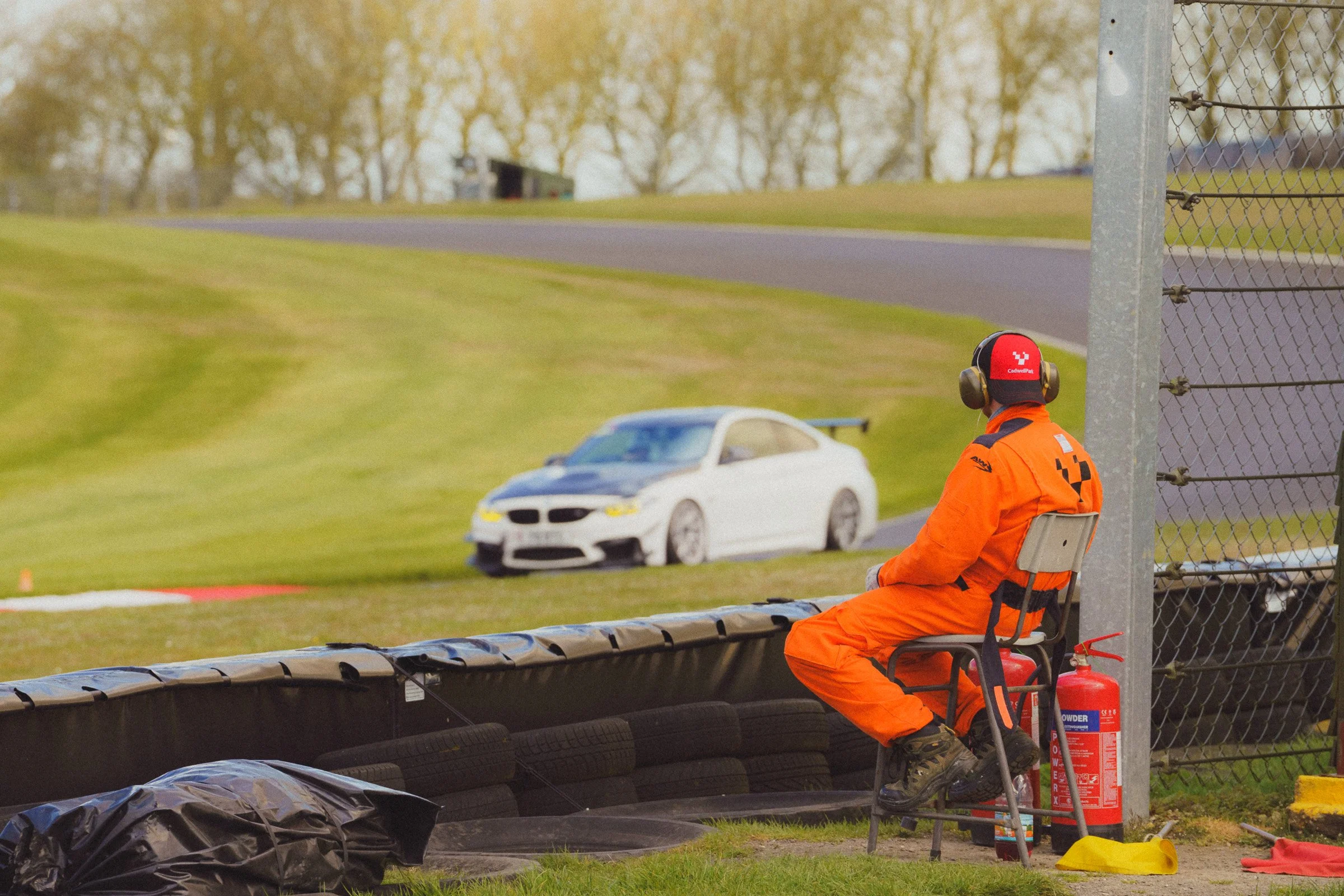 A race track scene with a track marshal in orange safety gear sitting on a chair near the track, watching a white race car pass by