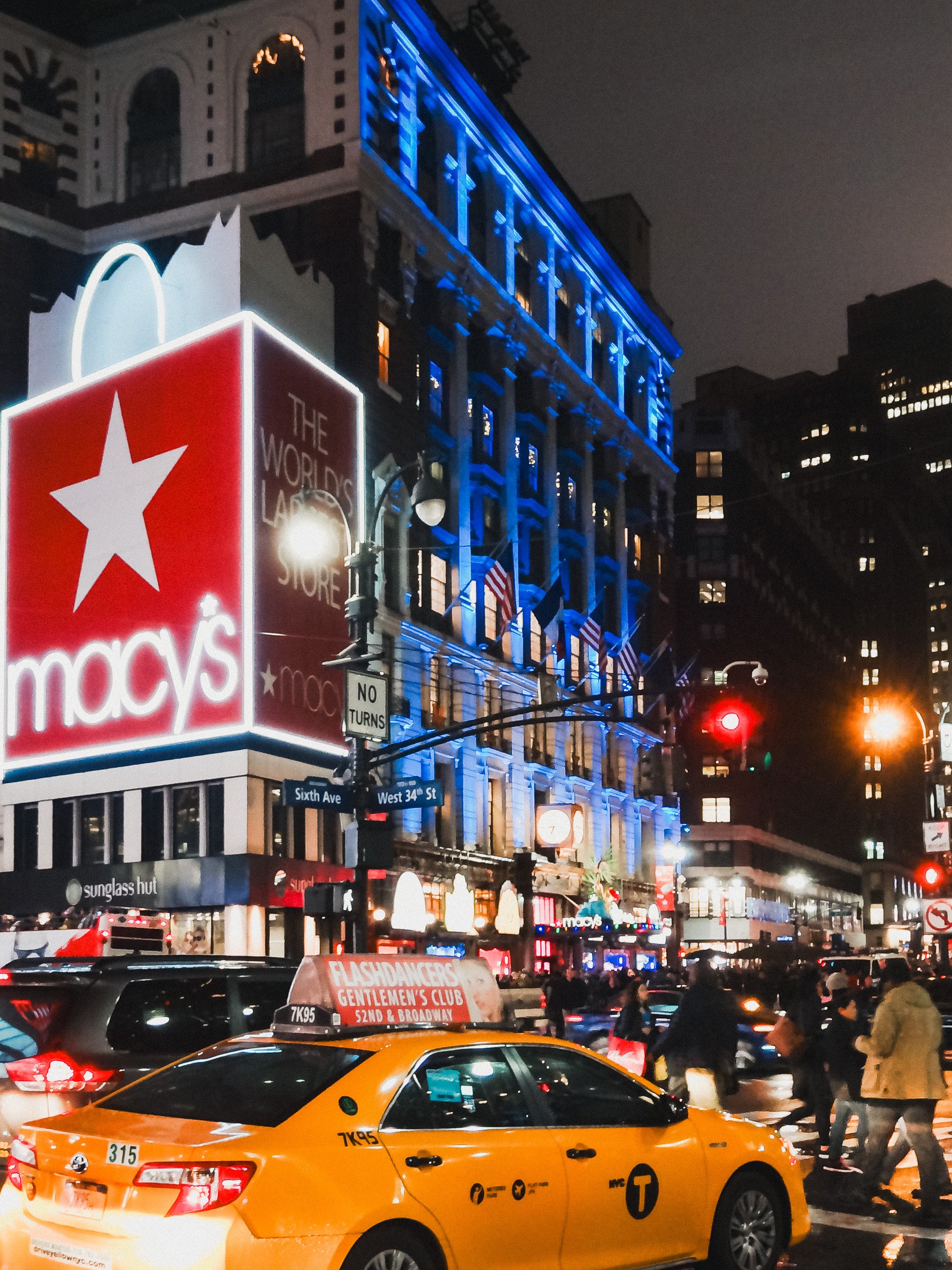 Night scene of Times Square in New York City, illuminated with bright neon signs, a Macy's sign, and a yellow taxi in the foreground with pedestrians crossing the street.
