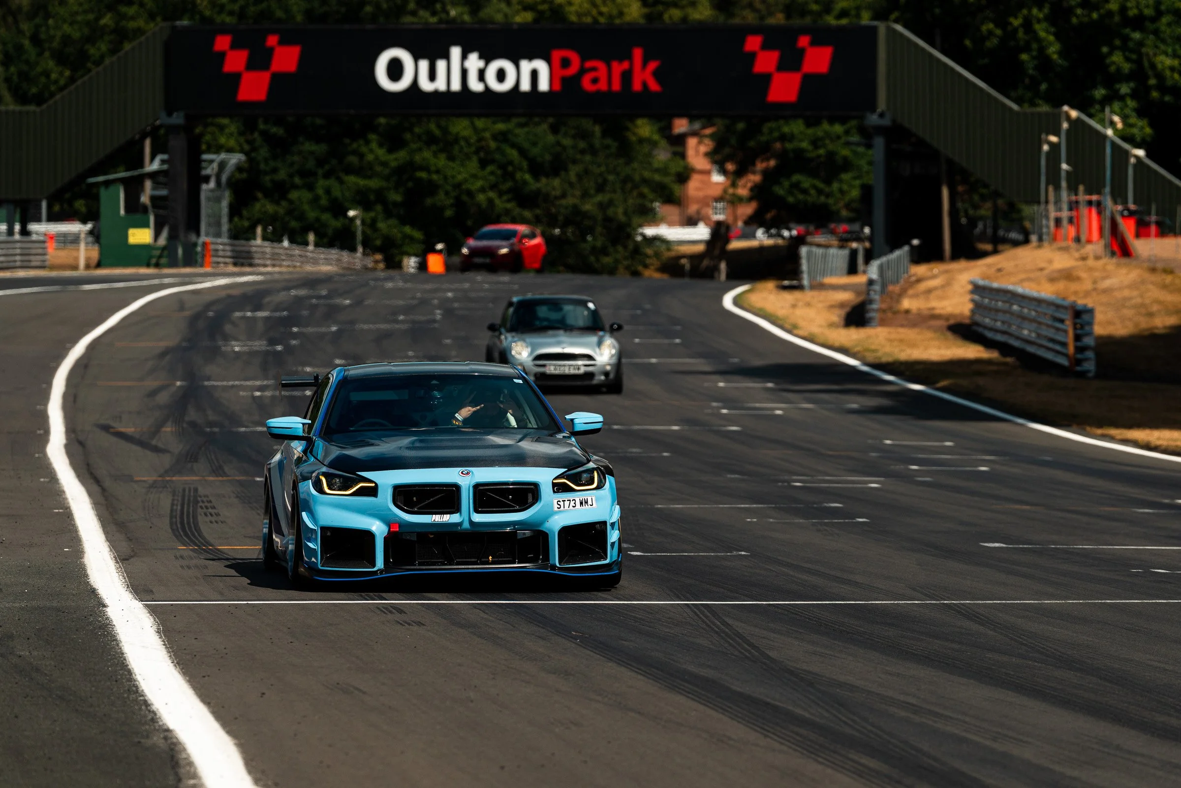 A bright blue sports car driving on a race track with a black and red sign overhead reading "Oulton Park," surrounded by green trees and a clear sky.