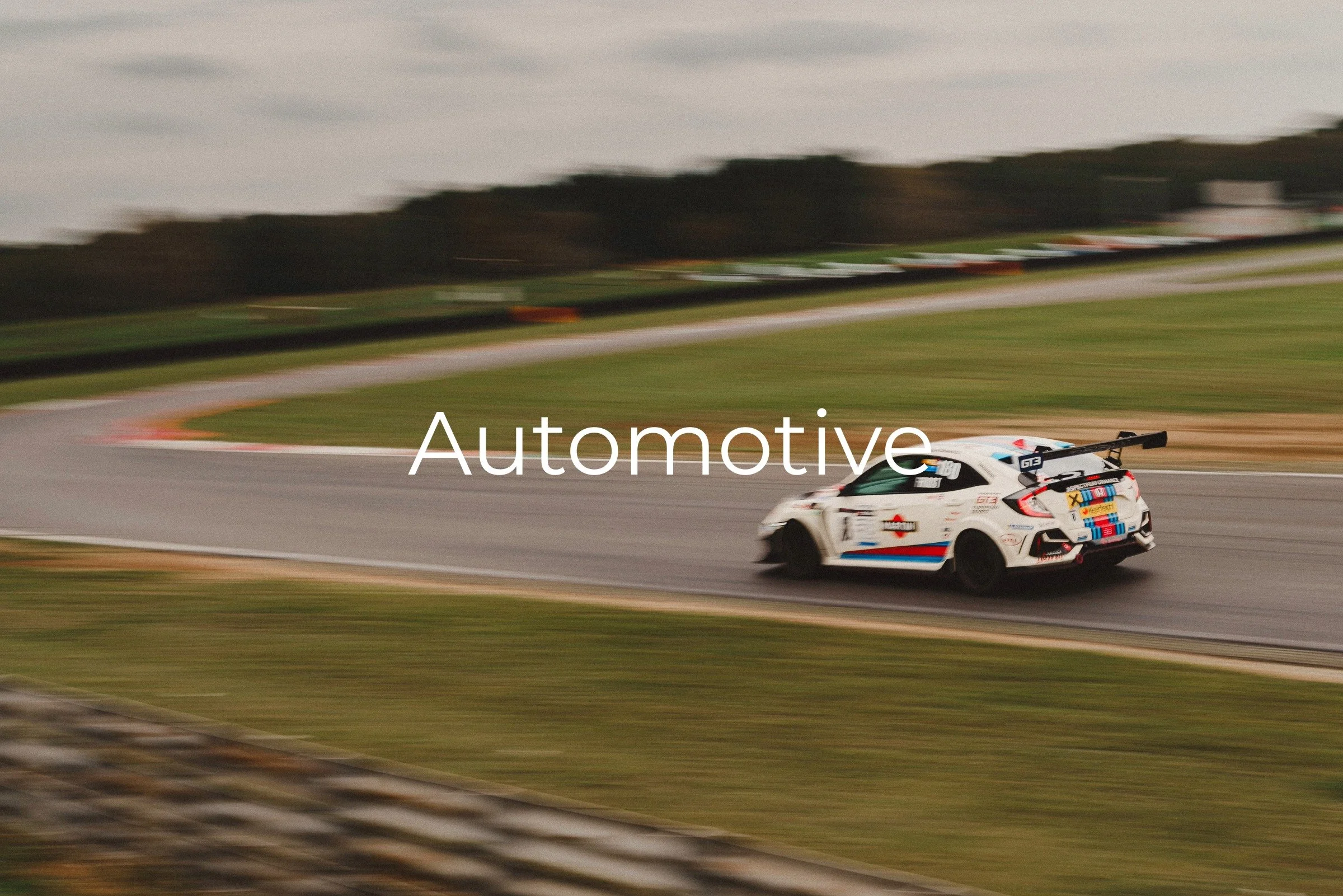 A white race car with colorful stripes speeds around a curve on a race track during daytime, with blurred background of trees and sky which indicates motion.