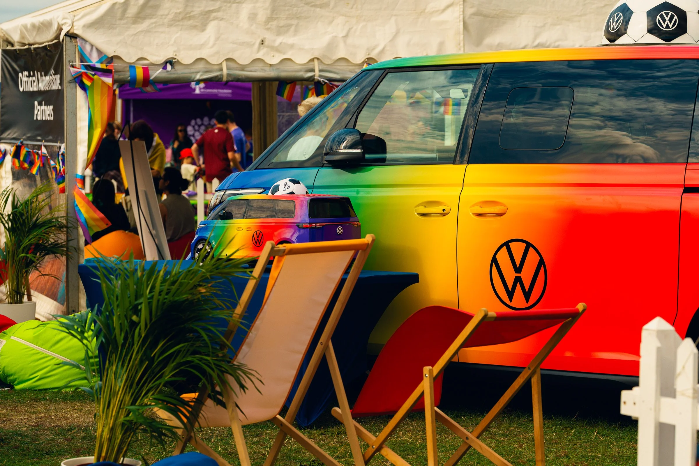 A Volkswagen van painted in rainbow colors parked at an outdoor pride event, with rainbow flags and people in the background.