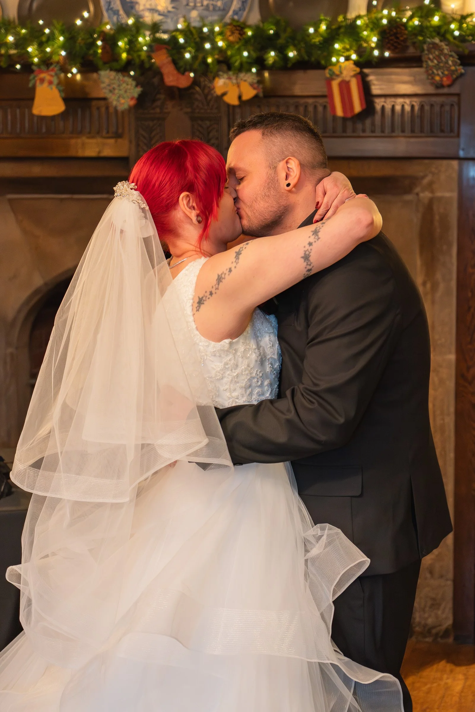 A bride with red hair and tattoos, wearing a white wedding dress and veil, and a groom in a black suit, sharing a kiss in front of a fireplace decorated with Christmas garland and stockings.