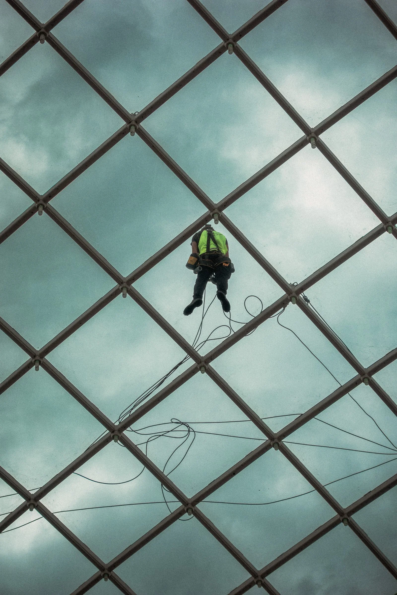 A worker in safety gear is suspended from a harness, working on a metal structure under a cloudy sky with a grid pattern overhead.