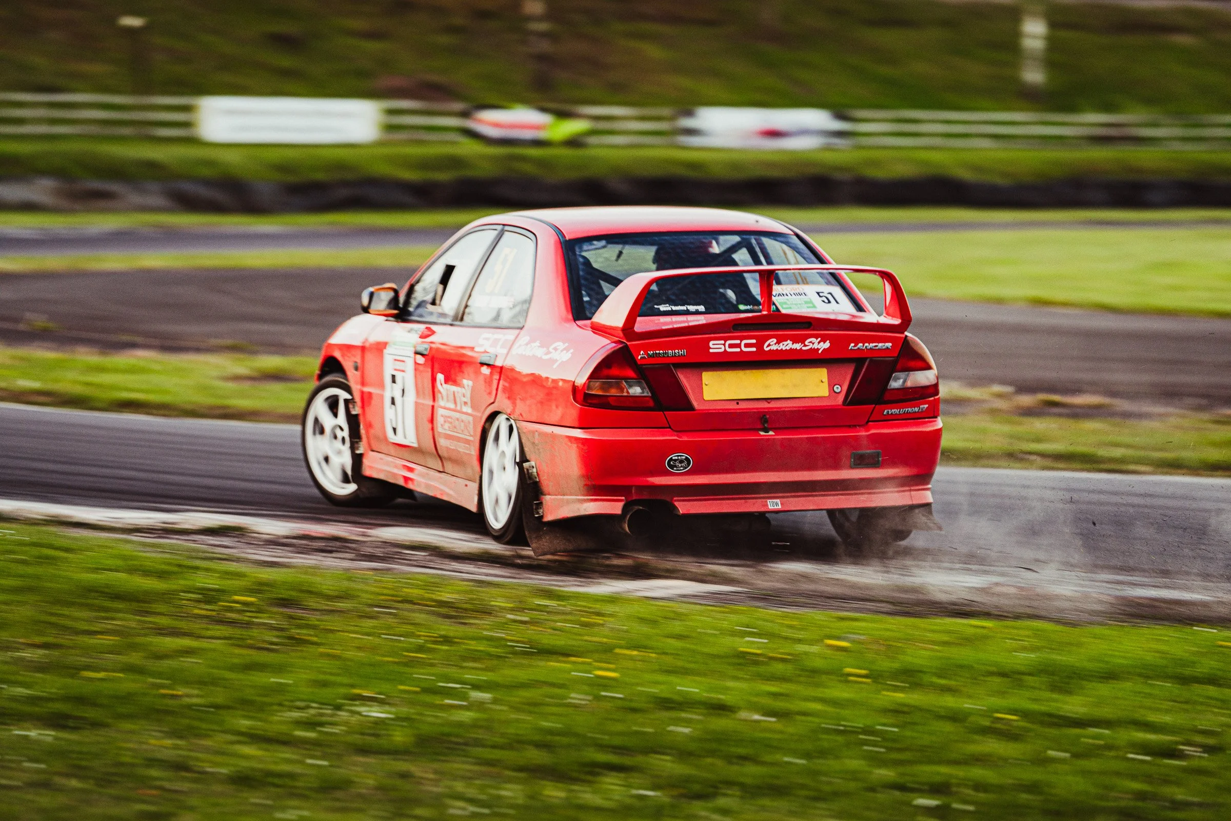 Red race car on a wet track, moving at high speed with a blurred background and grass on the sides.