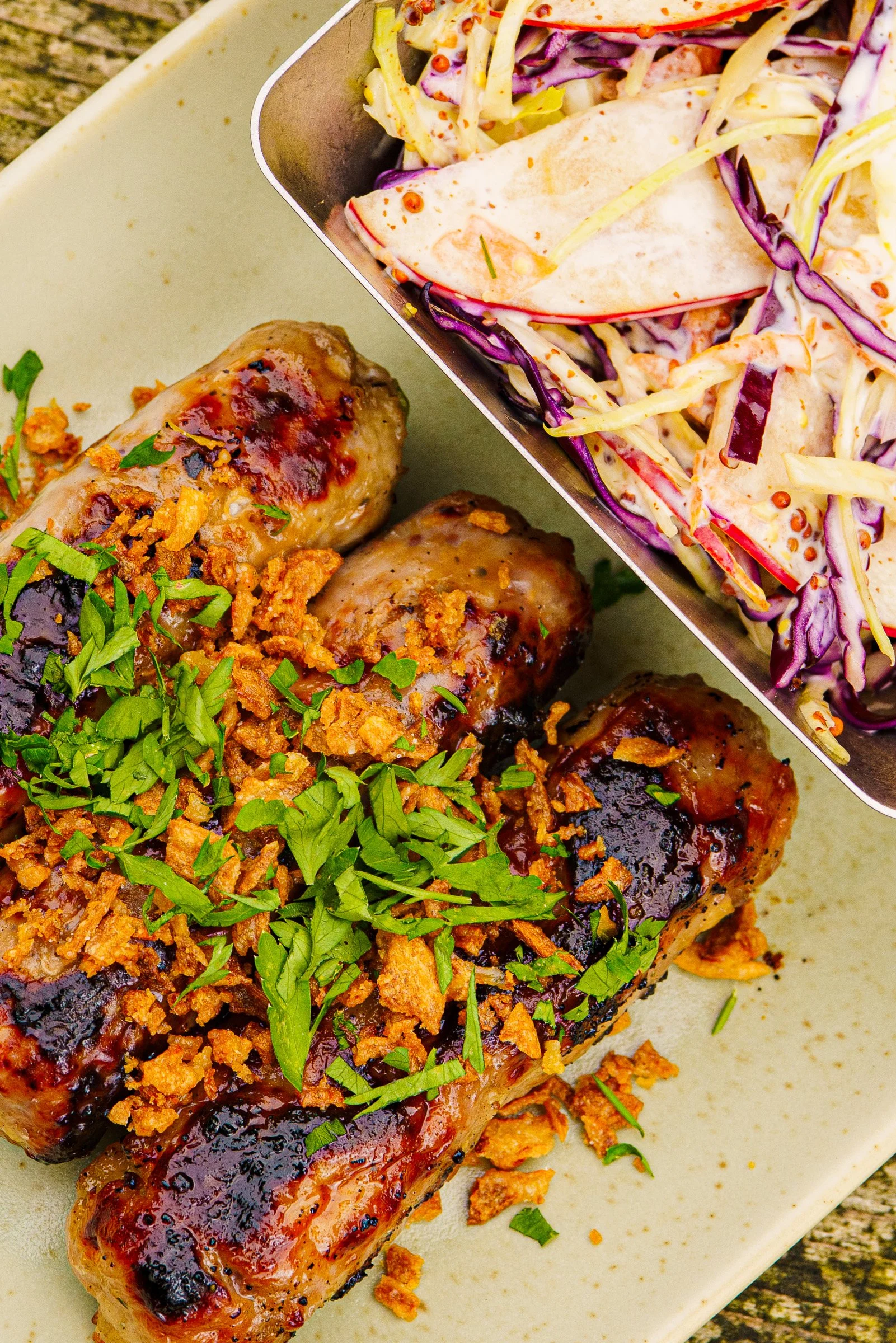 Grilled sausages, garnished with chopped green herbs and crispy fried onions, served on a light-coloured plate.