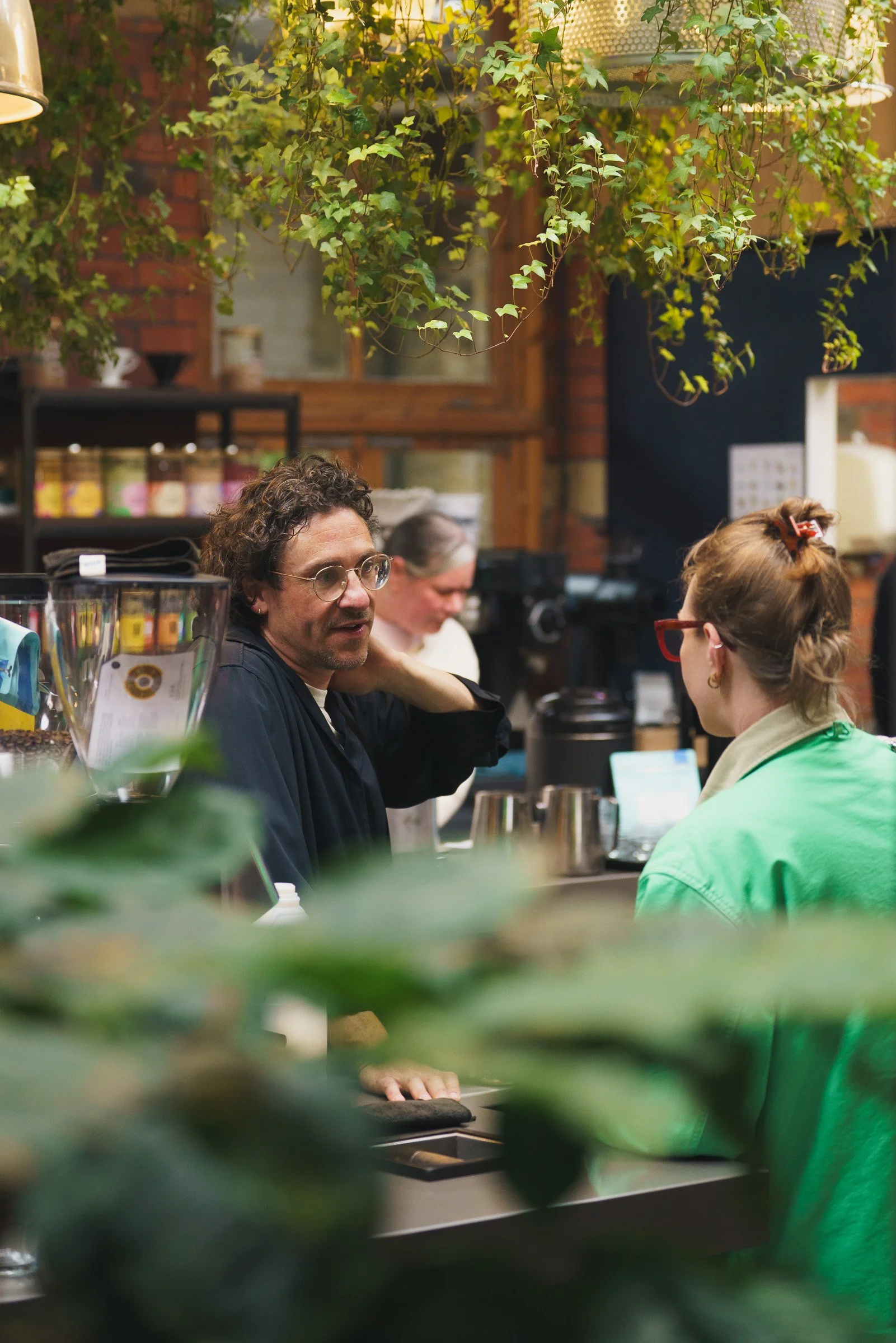 People sitting and talking at a counter in a cozy cafe with hanging greenery and warm wooden decor.