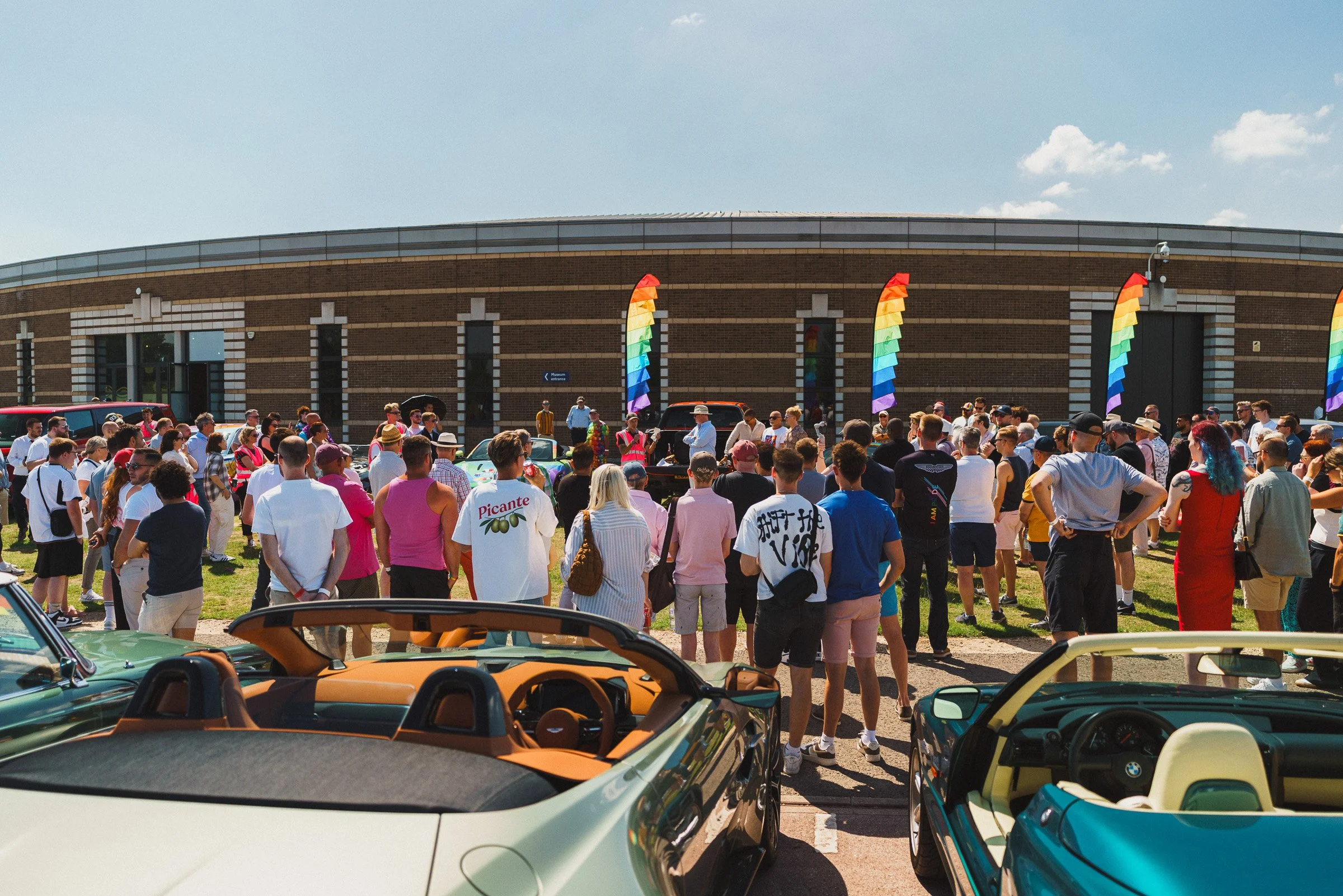A large group of people gathered outside in front of a brick building decorated with rainbow flags.