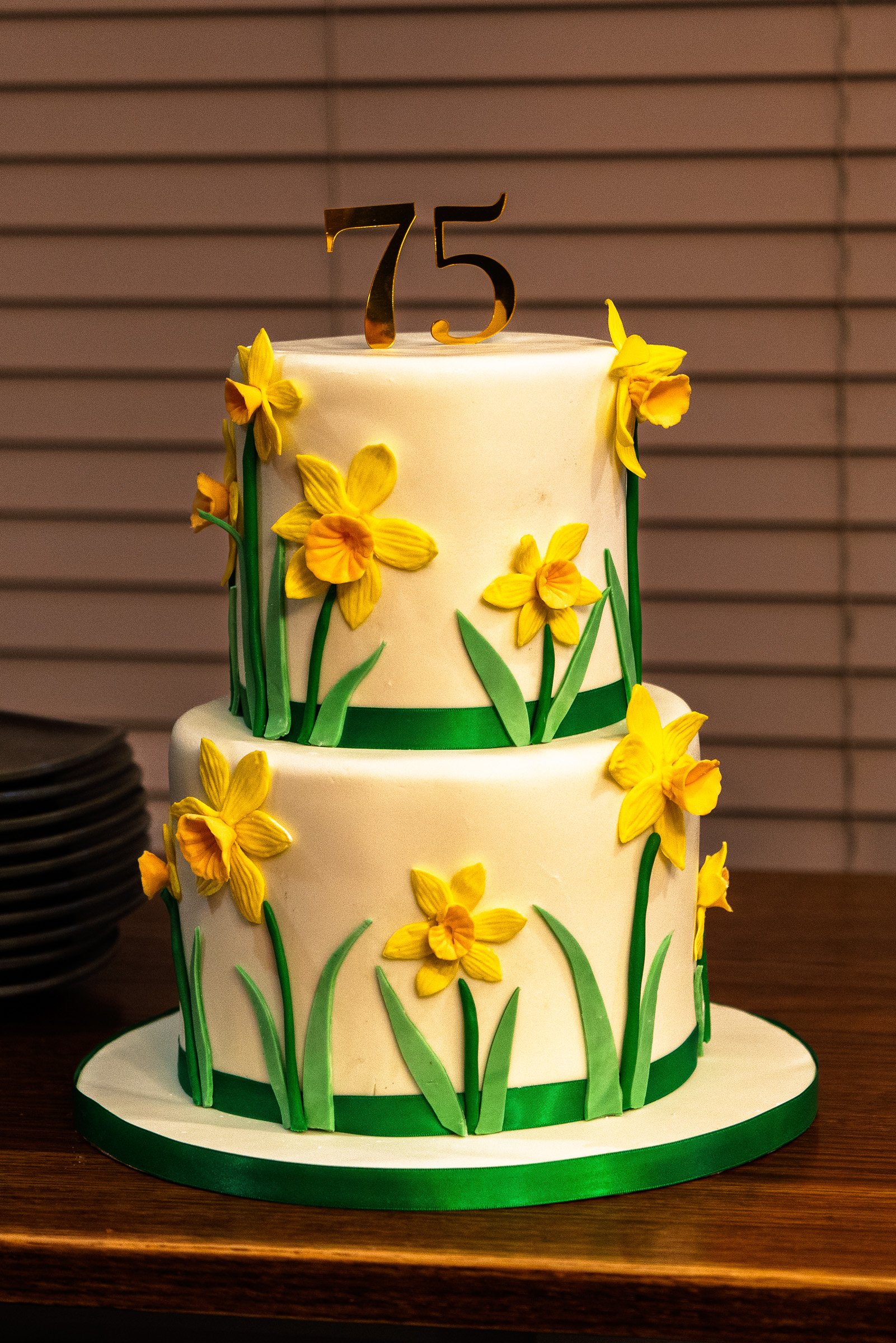 A two-tiered white cake decorated with yellow flowers and green grass-like accents. A gold topper with the number '75' is on top of the cake.