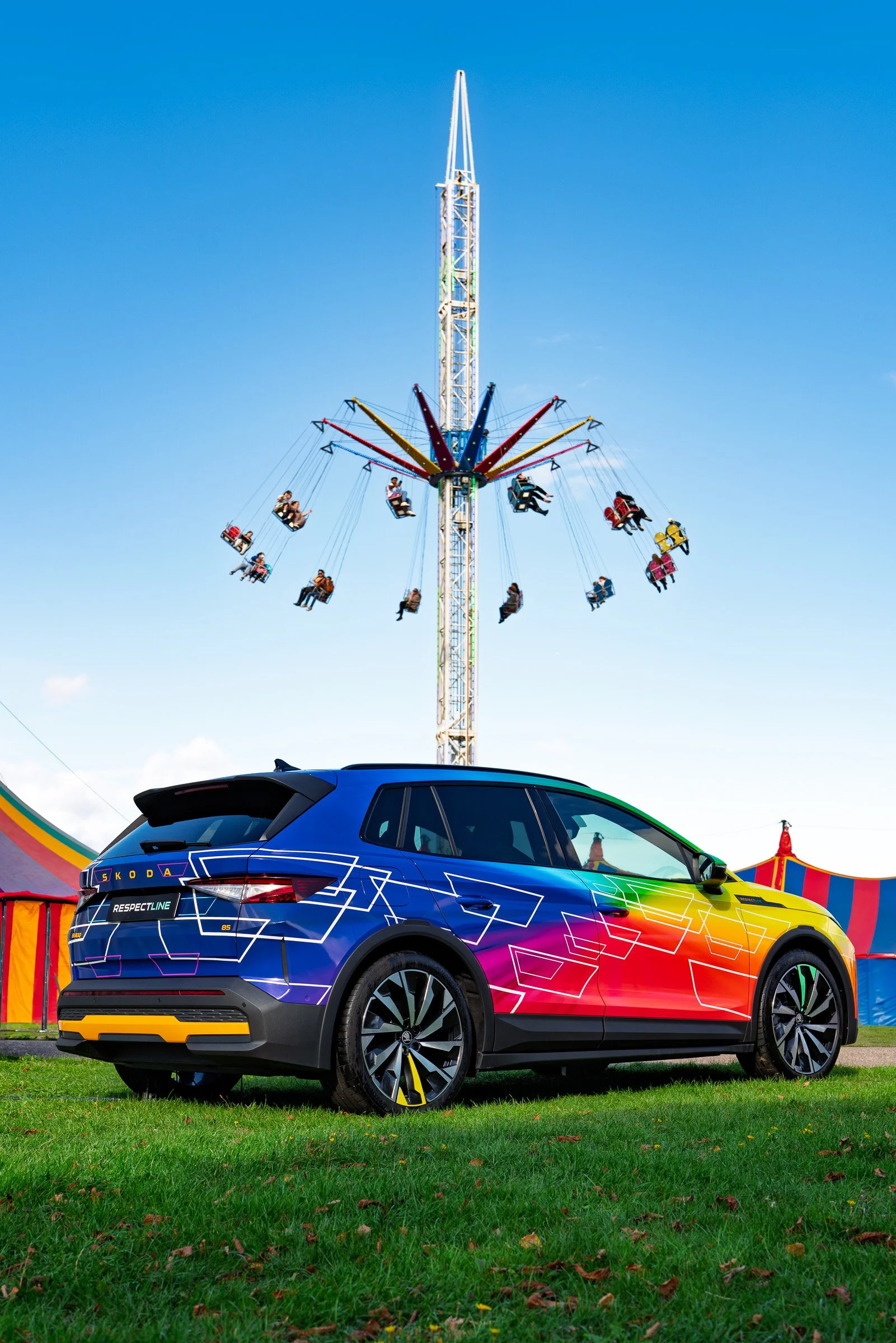 Colourful car parked on grass with a carnival swing ride in the background under a blue sky.