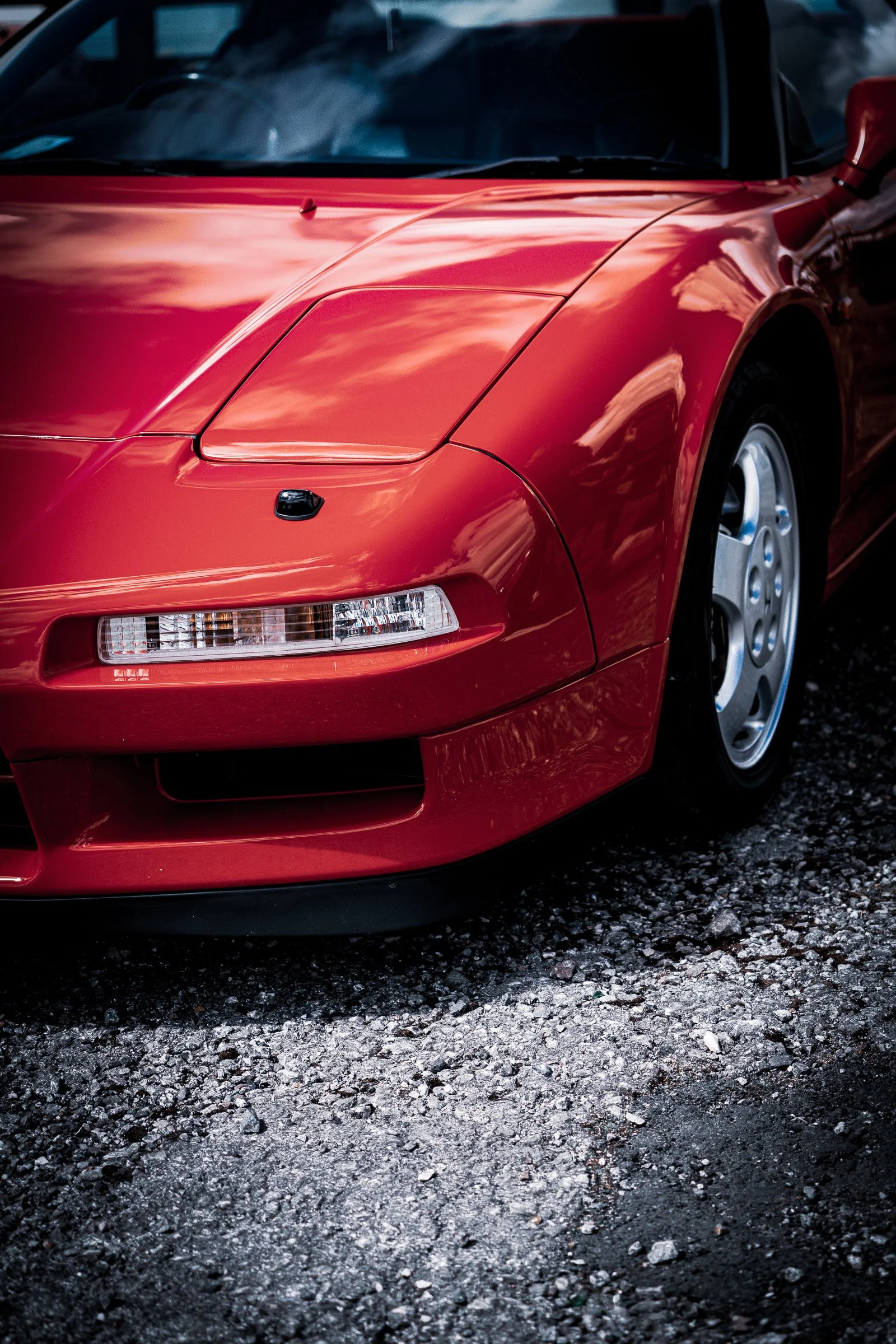 Close-up of a red sports car with sleek curves, parked on gravel, reflecting a cloudy sky on its shiny surface.