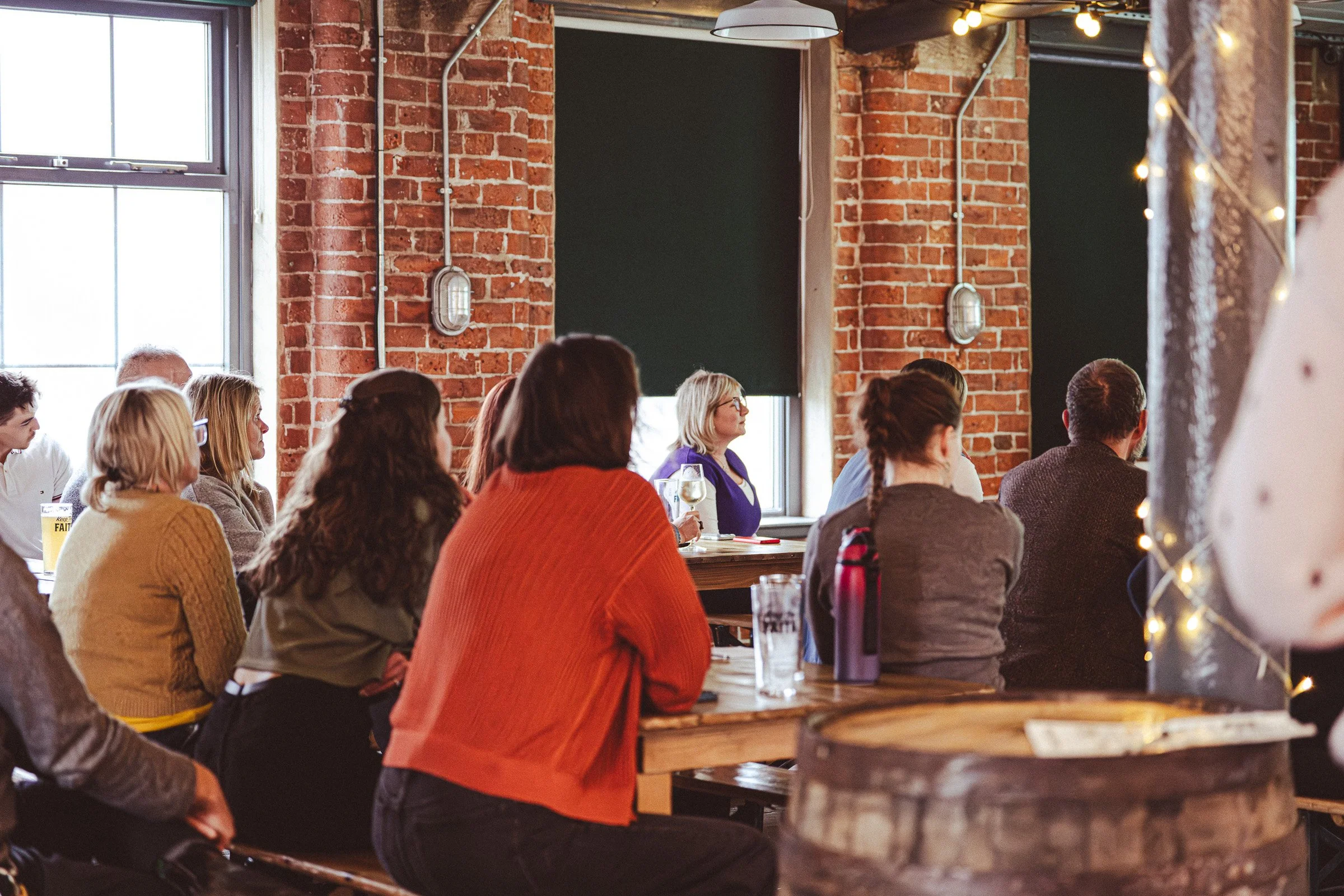 A group of people sitting on benches and at a long wooden table, attentively watching a presentation in a rustic brick-walled space with large windows and string lights.