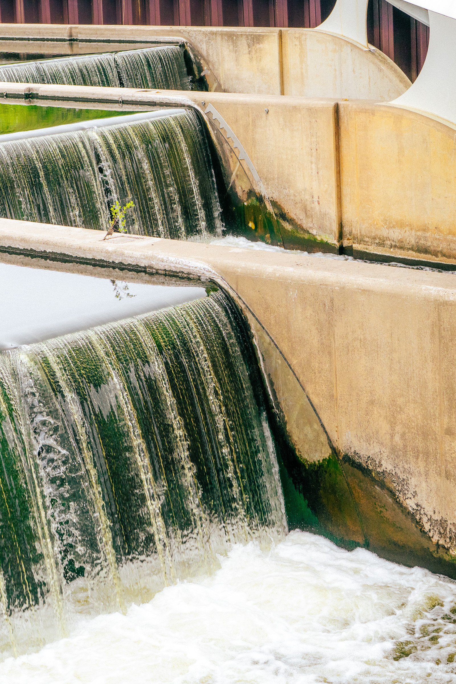 Water flows over multiple shallow concrete dams in a river, creating small waterfalls and white foam at the bottom.