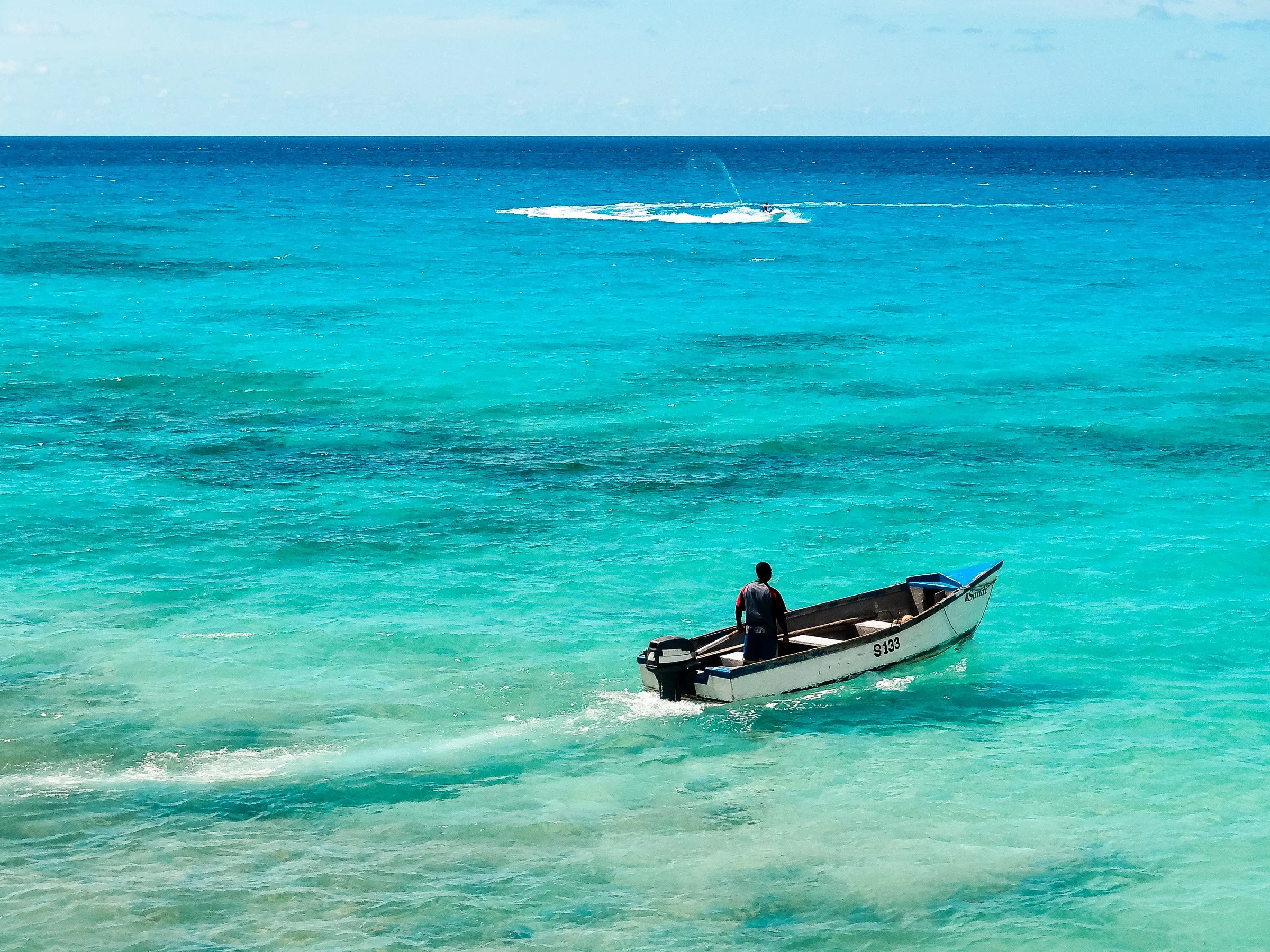 A boat with a person standing in it on bright turquoise water near the sandy beach and another person riding a jet ski in the distance on the ocean.