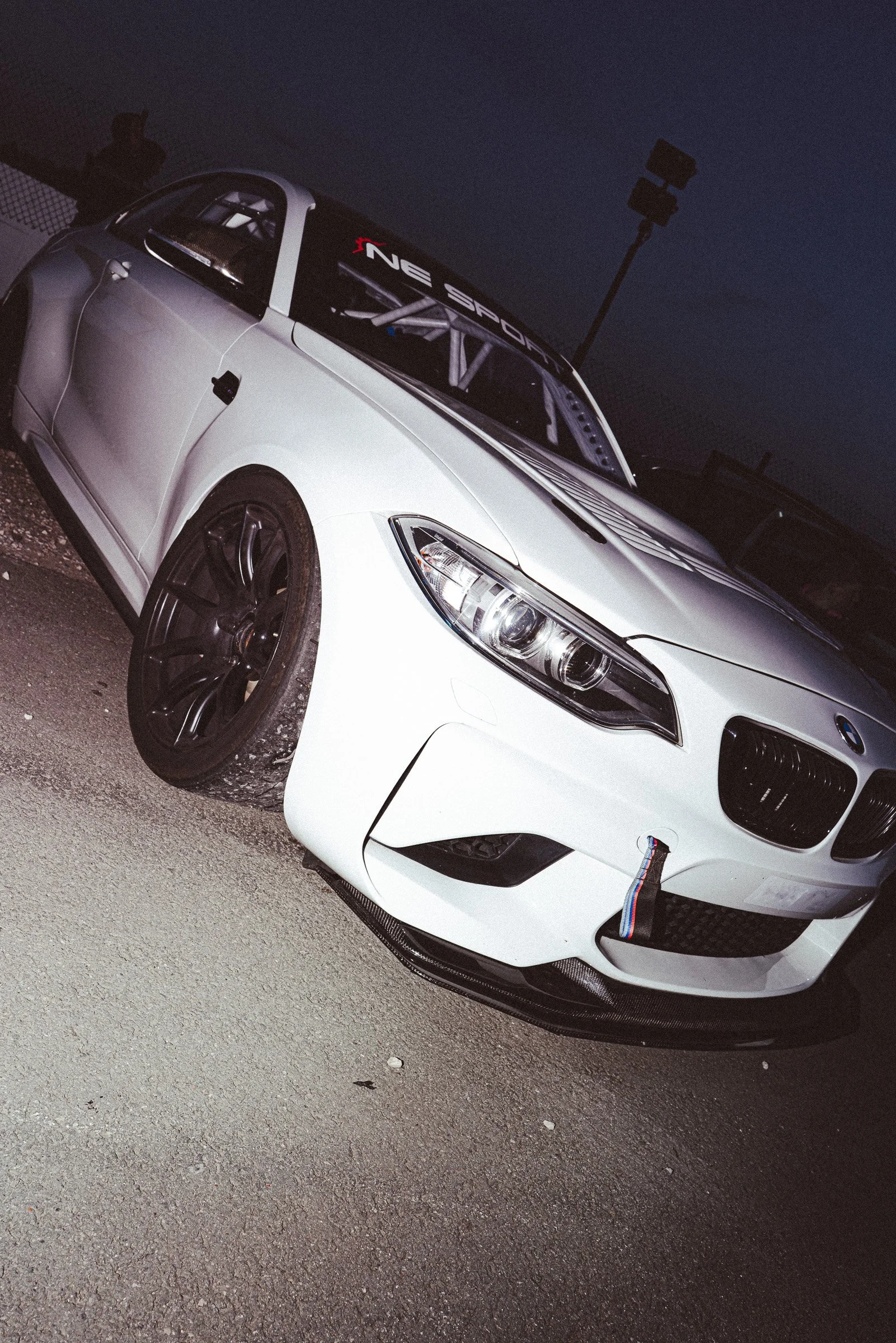 A white BMW M2 sports car parked on a gravel surface at night, with a racing roll cage visible inside. The car has black wheels and a low, aerodynamic design.