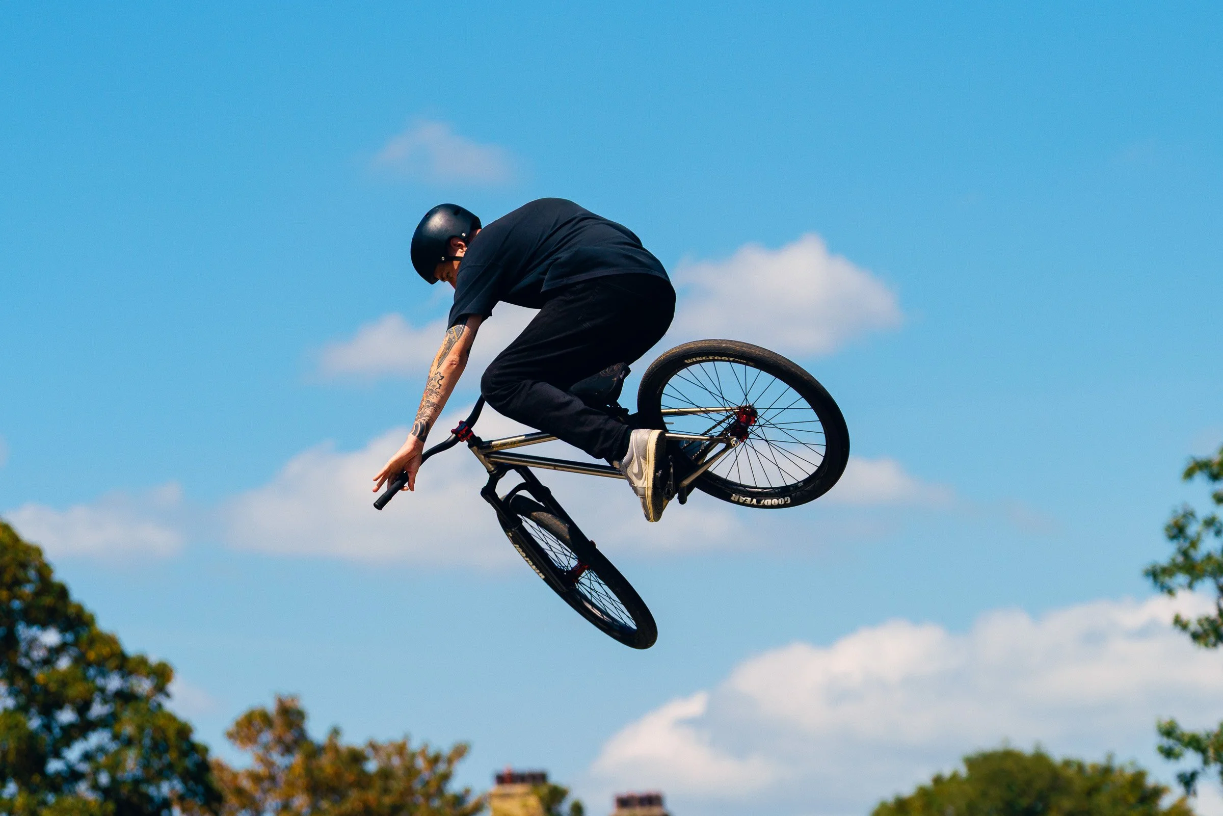 A person wearing a black helmet and dark clothing performing a stunt on a BMX bike in mid-air against a blue sky with some clouds.