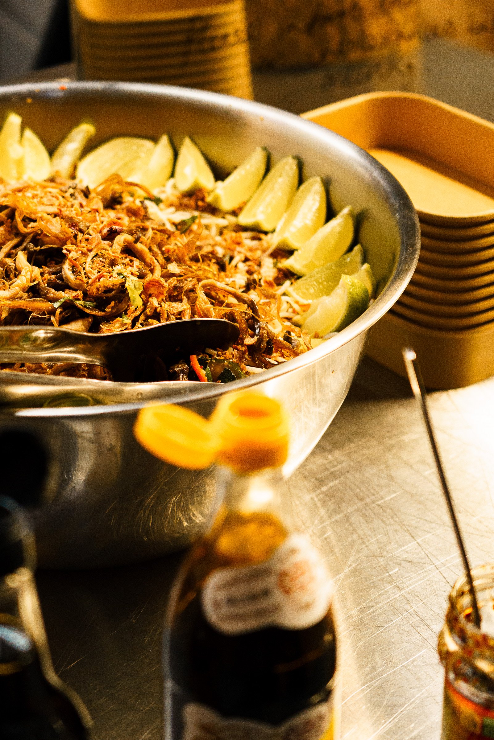 A large bowl of Thai noodle salad, garnished with lime wedges, with bottles of hot sauce and seasoning nearby.