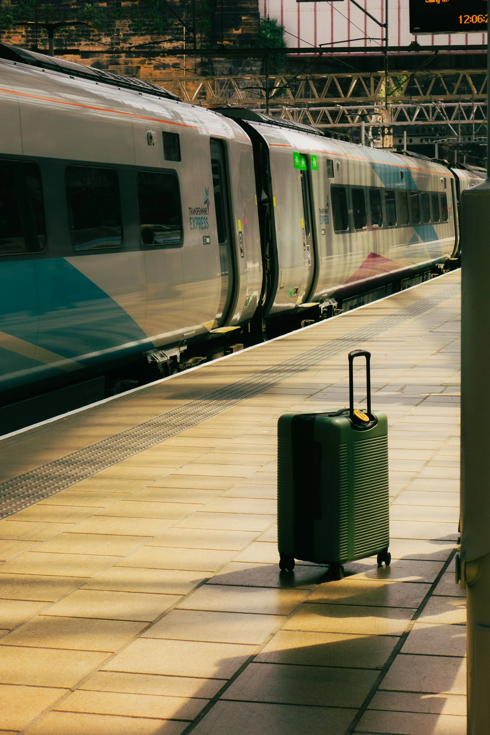 A silver passenger train at a platform with a green wheeled suitcase nearby, and some overhead signs and infrastructure in the background.