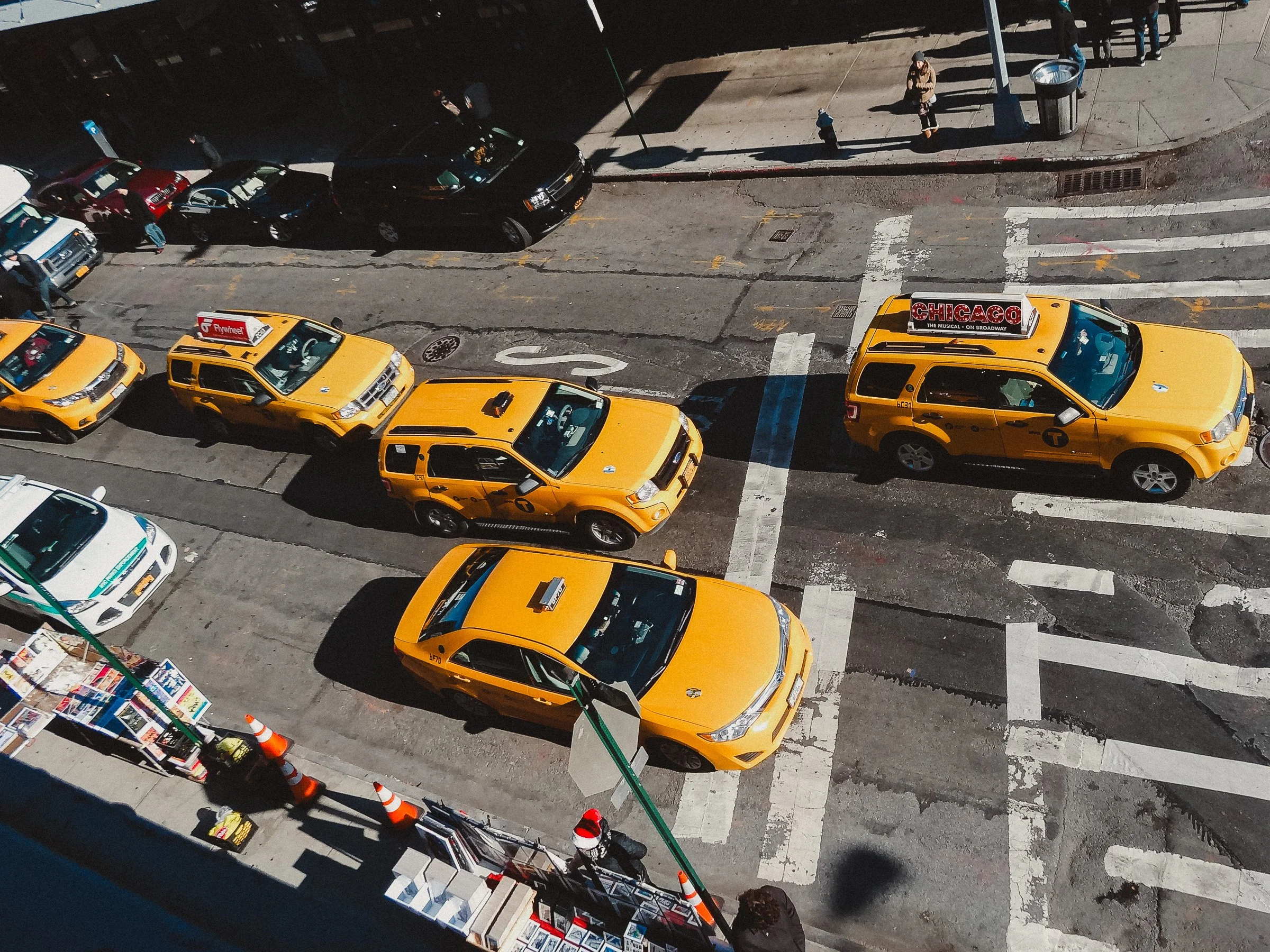 Bird's-eye view of a city street with yellow taxis, parked cars, pedestrians, a sidewalk vendor cart, and a newsstand in the lower left corner.