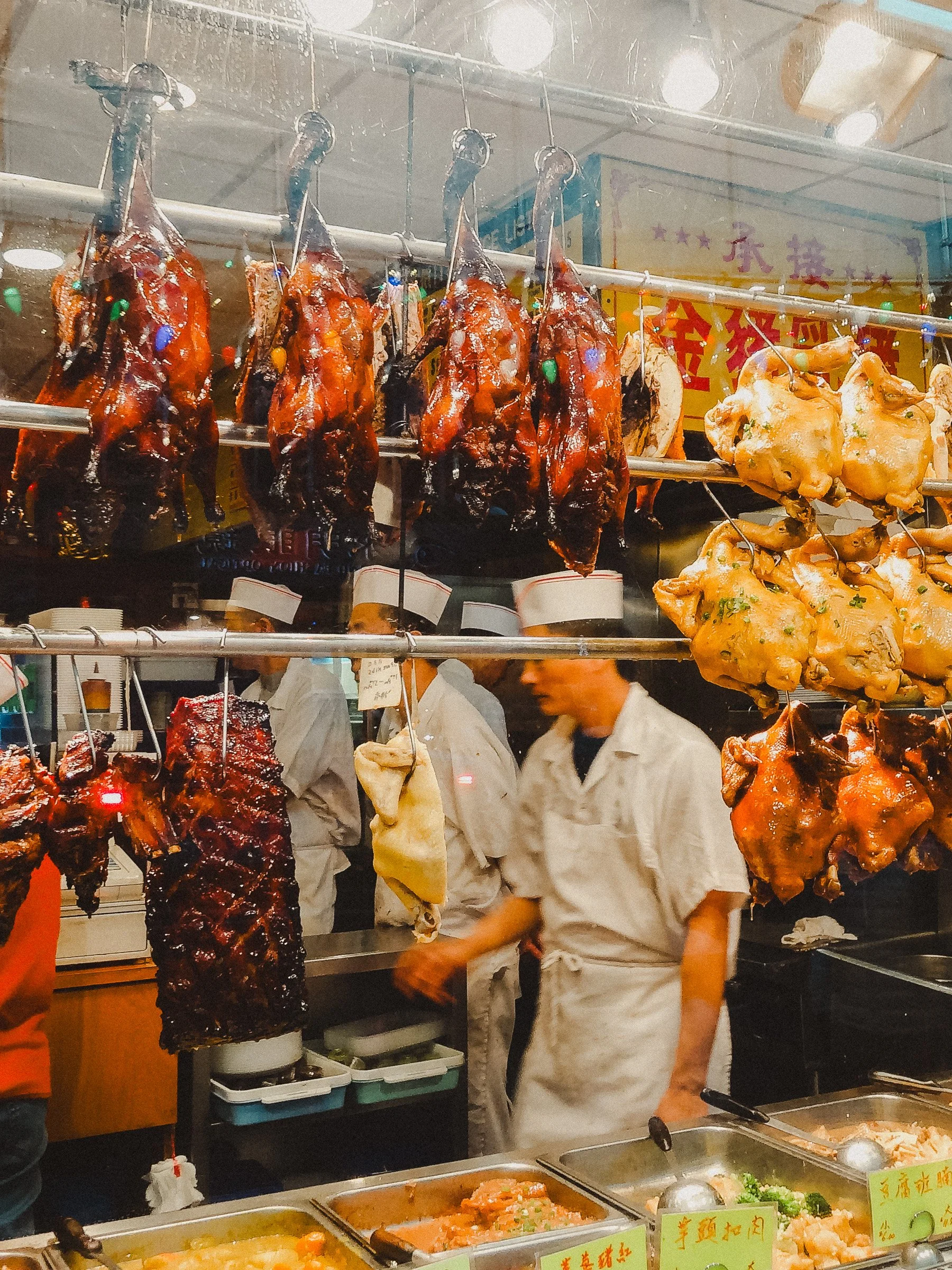 Hanging roasted ducks and chickens at a street food stall in an Asian market, with food containers in front and workers preparing in the background.