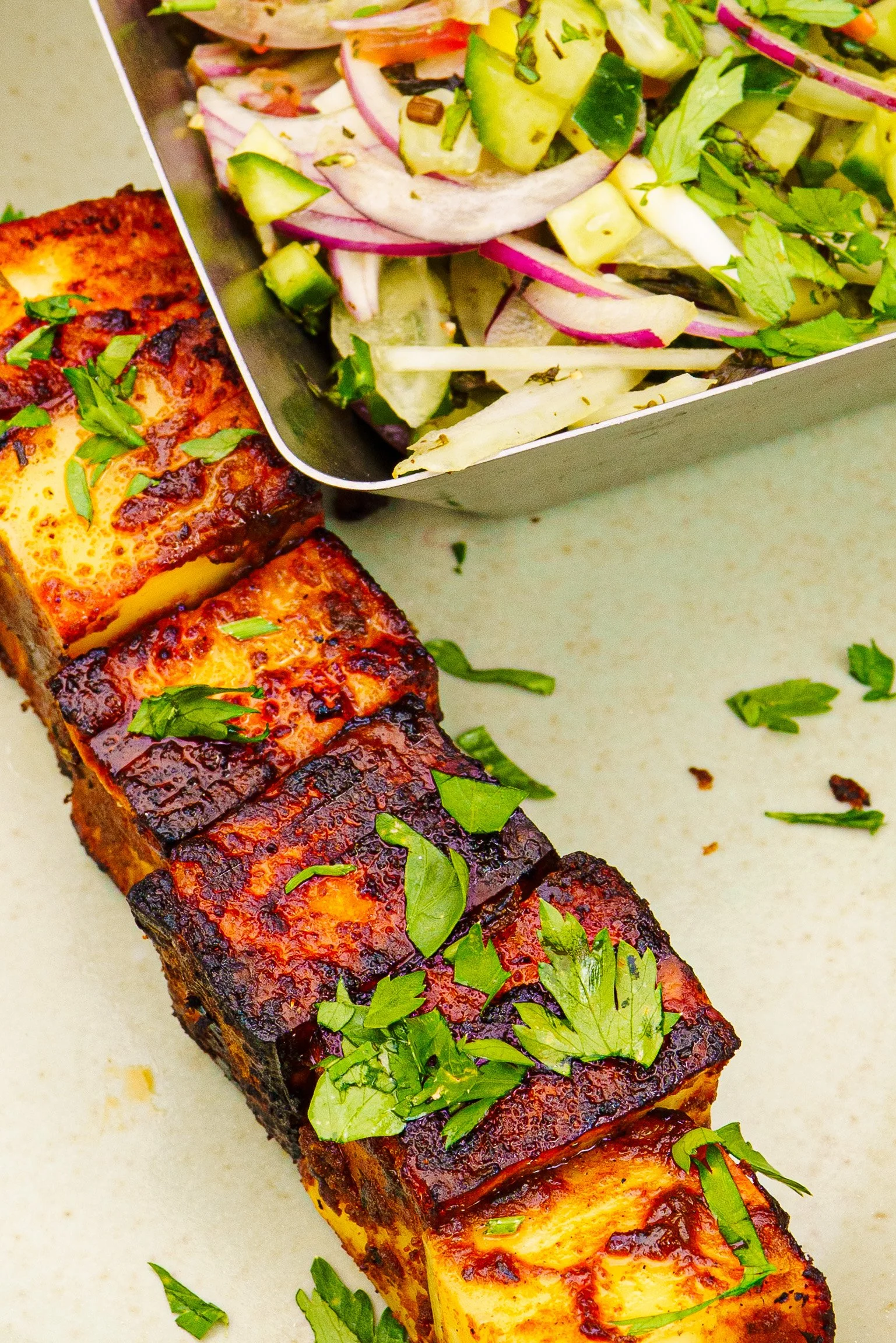 Close-up of grilled paneer cheese with a dark char and herbs on top, with a side salad garnished with chopped fresh herbs, on a light-coloured surface.