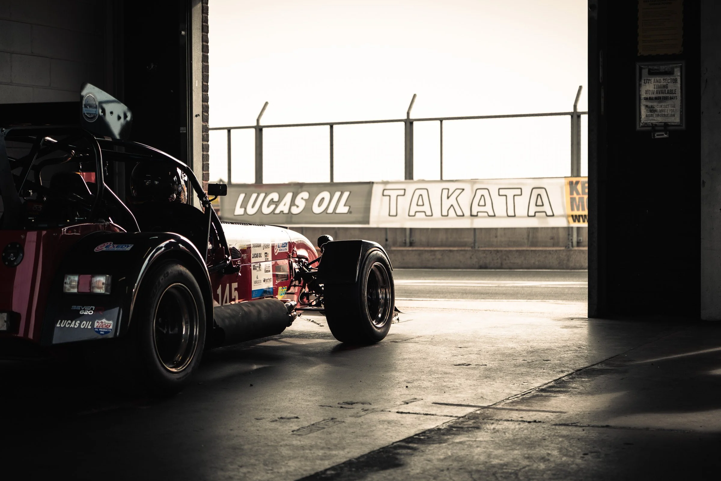 A race car is inside a garage, partially visible with decals and stickers, with the garage door open revealing a track with banners and fencing outside.