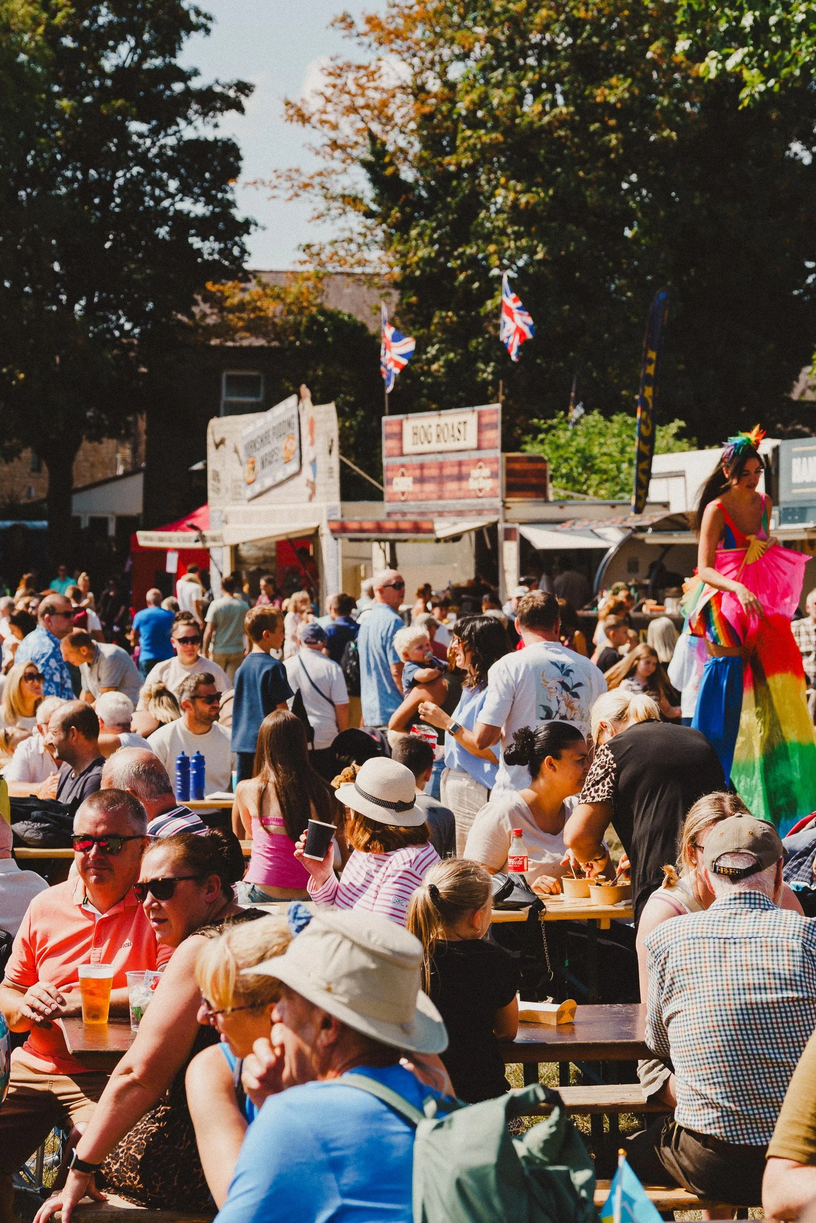 Crowd gathered outdoors at festival with food stalls, flags, and a performer in colorful attire.