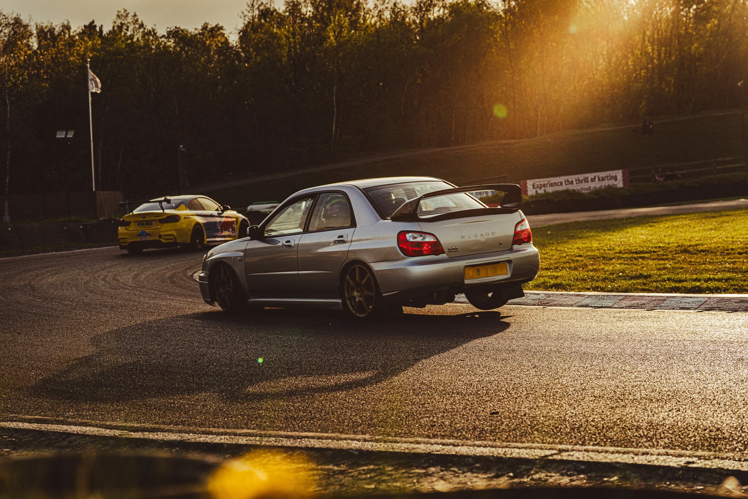 Two cars on a racetrack at sunset, with a wooded area in the background.