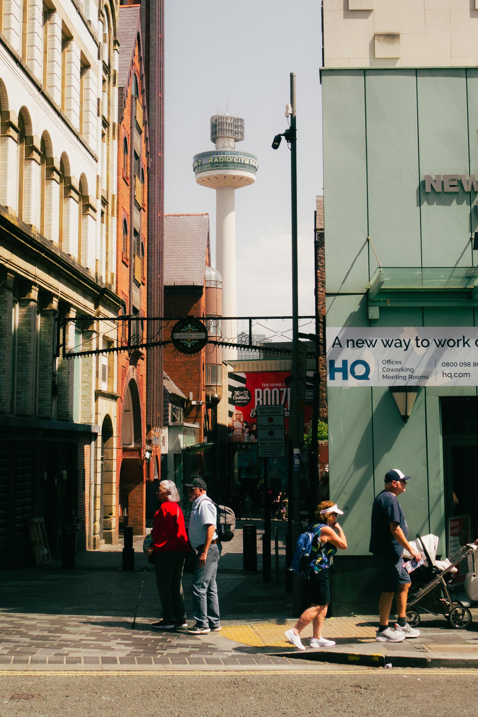 People walking on a city street with buildings and a tower featuring a television antenna in the background.