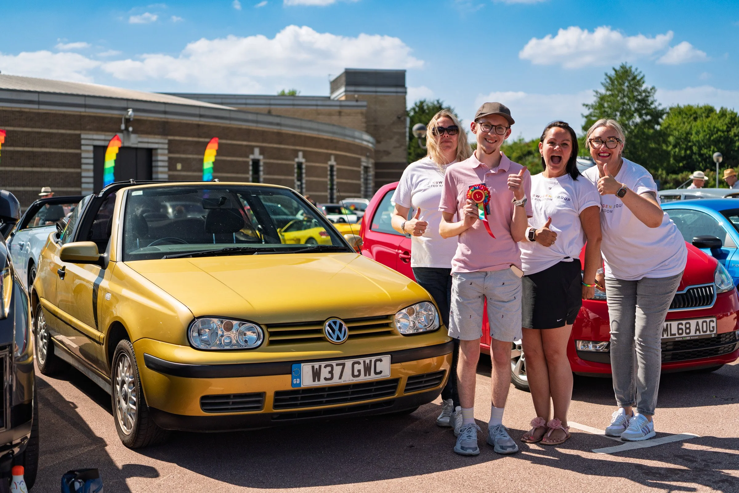 Group of four people smiling and celebrating, standing behind a yellow Volkswagen car at a pride-themed car show, with rainbow flags on a building in the background.
