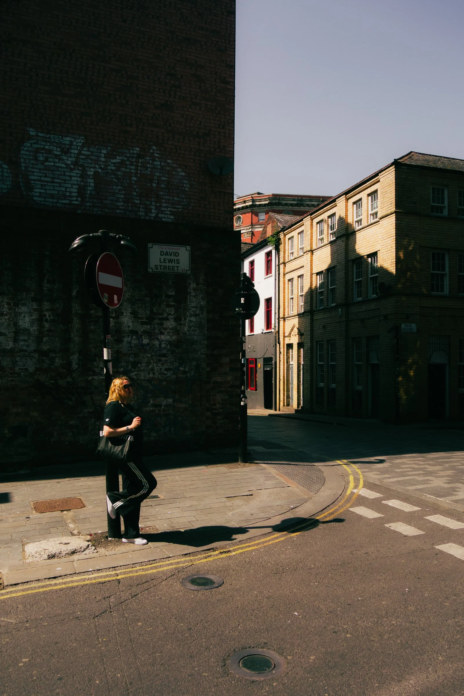 Street scene with a woman walking on a sidewalk, shadowed buildings in the background, street signs, and a street with painted lines and drainage covers.
