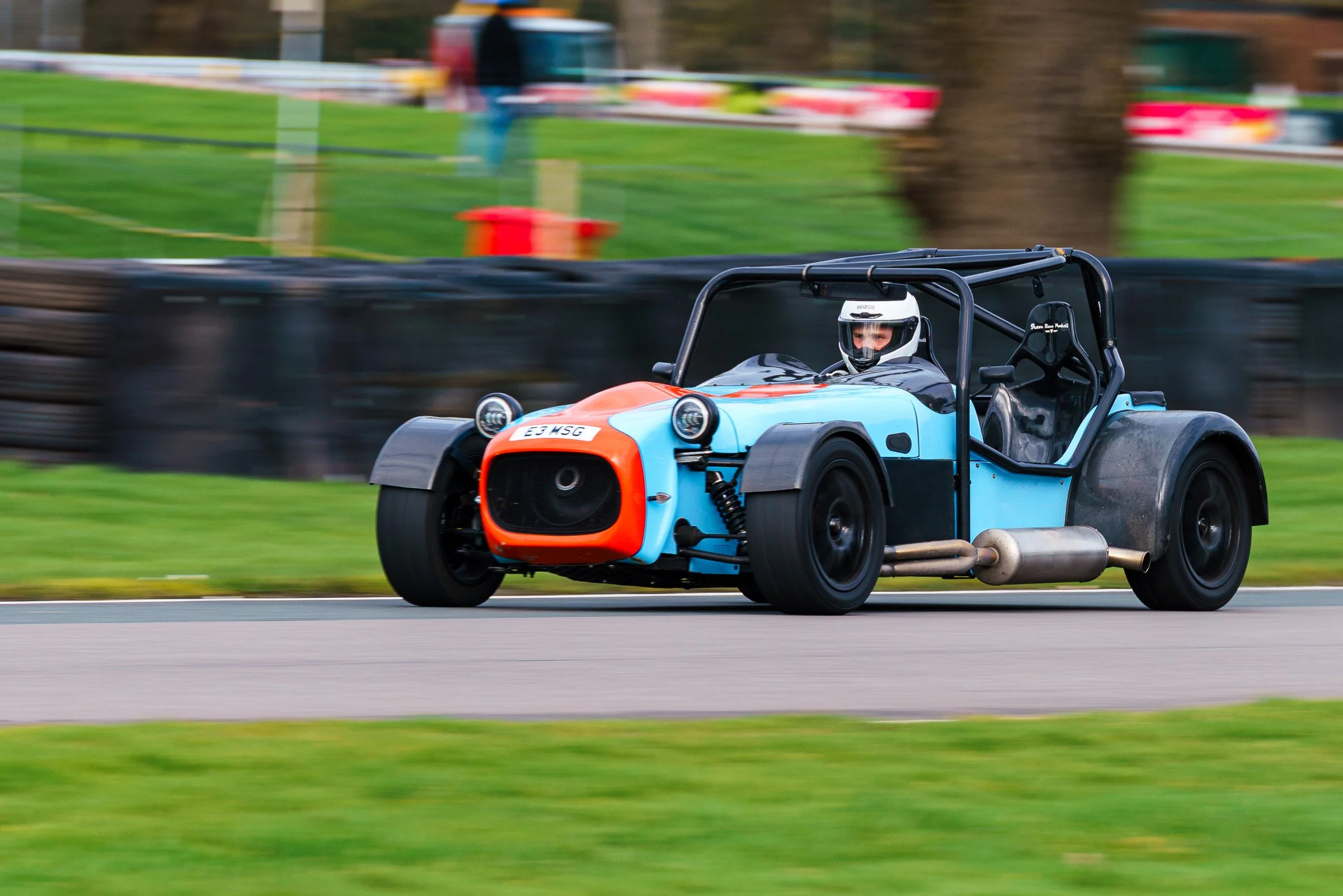 A race car with a blue and orange body speeding on a racetrack, with a driver wearing a helmet, surrounded by a blurred background of trees and barriers.