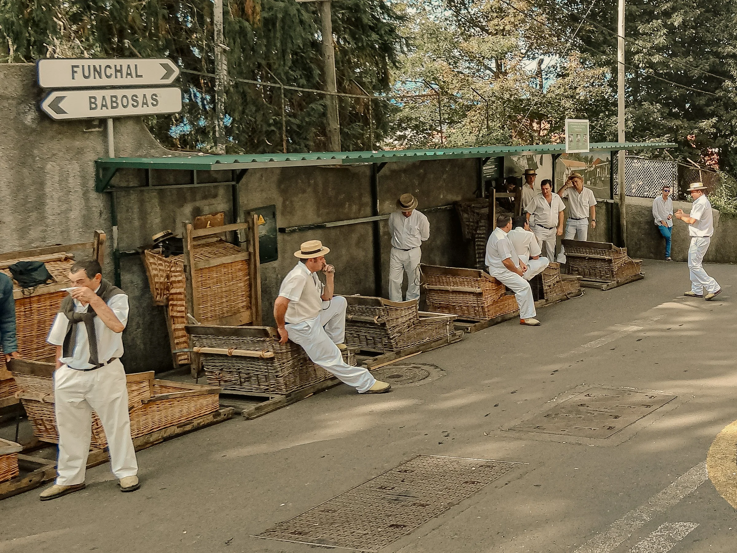 Carreiros do Monte dressed in white clothing and hats, some sitting and some standing, near a green-roofed structure and wicker sledges, on a street with trees in the background.