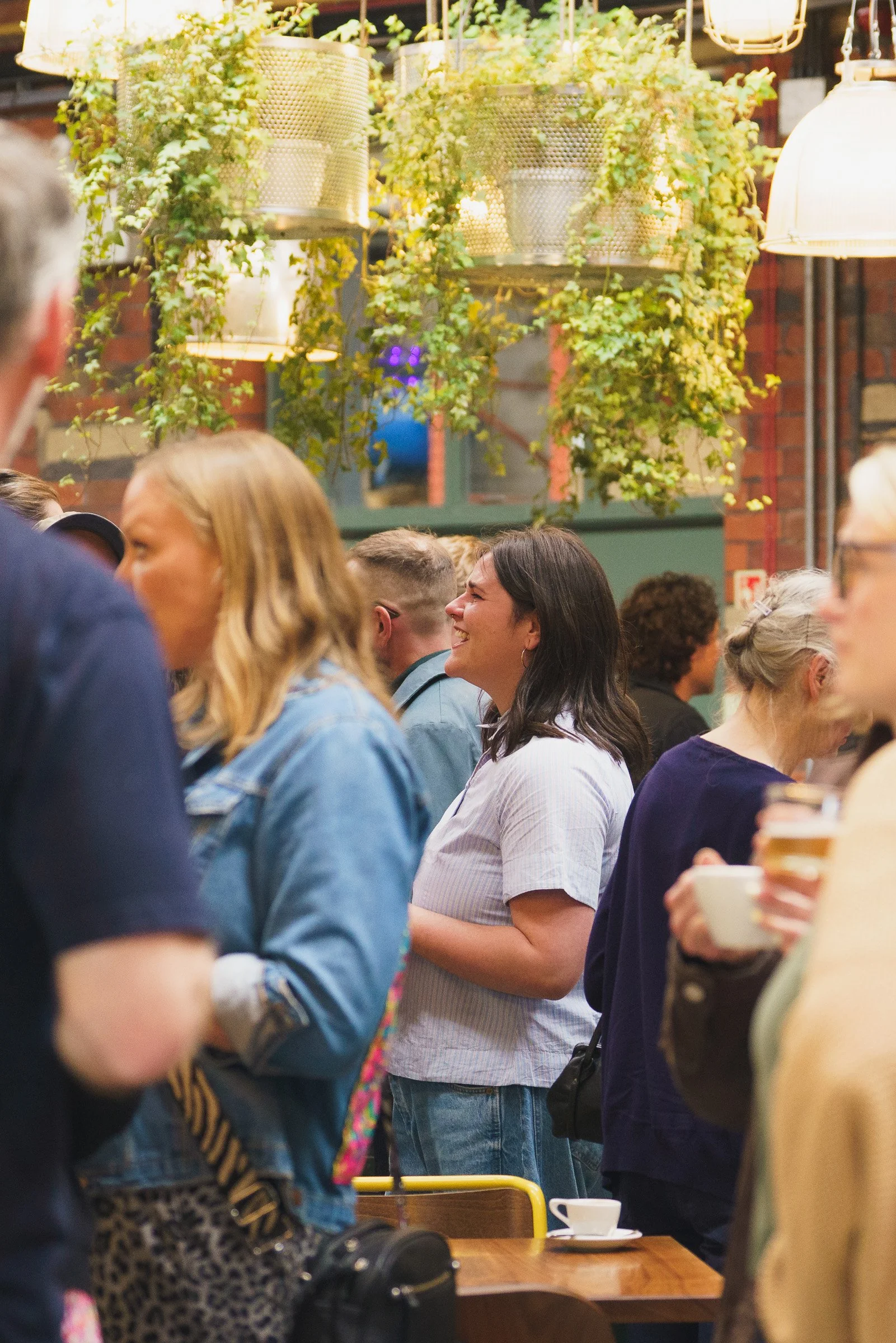 A group of people standing in line at a coffee shop, with hanging plants and warm lighting overhead.