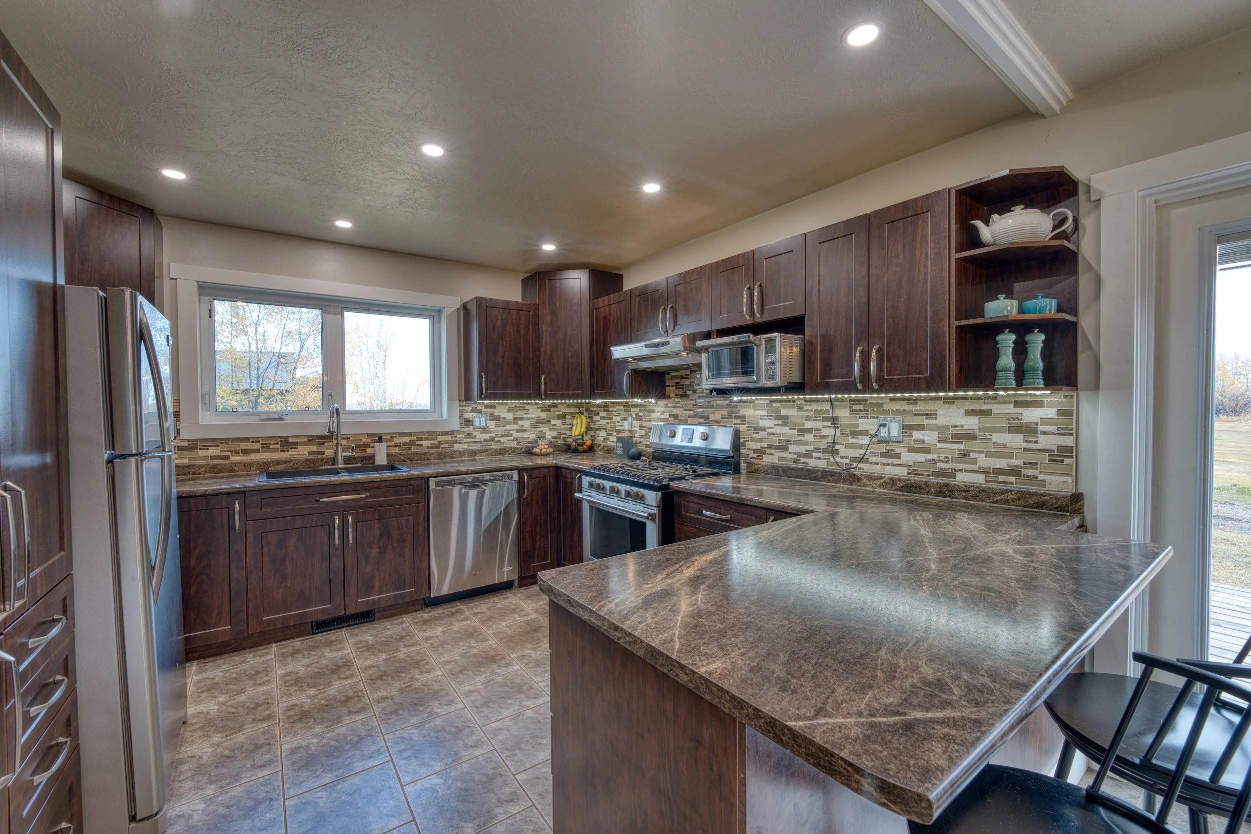 Bright, modern kitchen with white cabinets, a white island with a marble top, gold hardware and fixtures, pendant lights, and a dining area with a round table and woven chairs. Large windows and a glass door provide natural light.