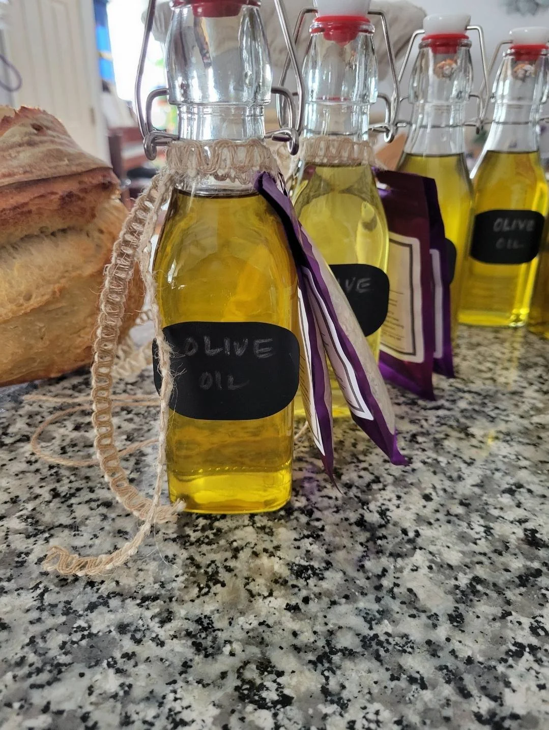 Bottles of olive oil with black labels and purple and white tags on a granite countertop, with a loaf of bread partially visible on the left.