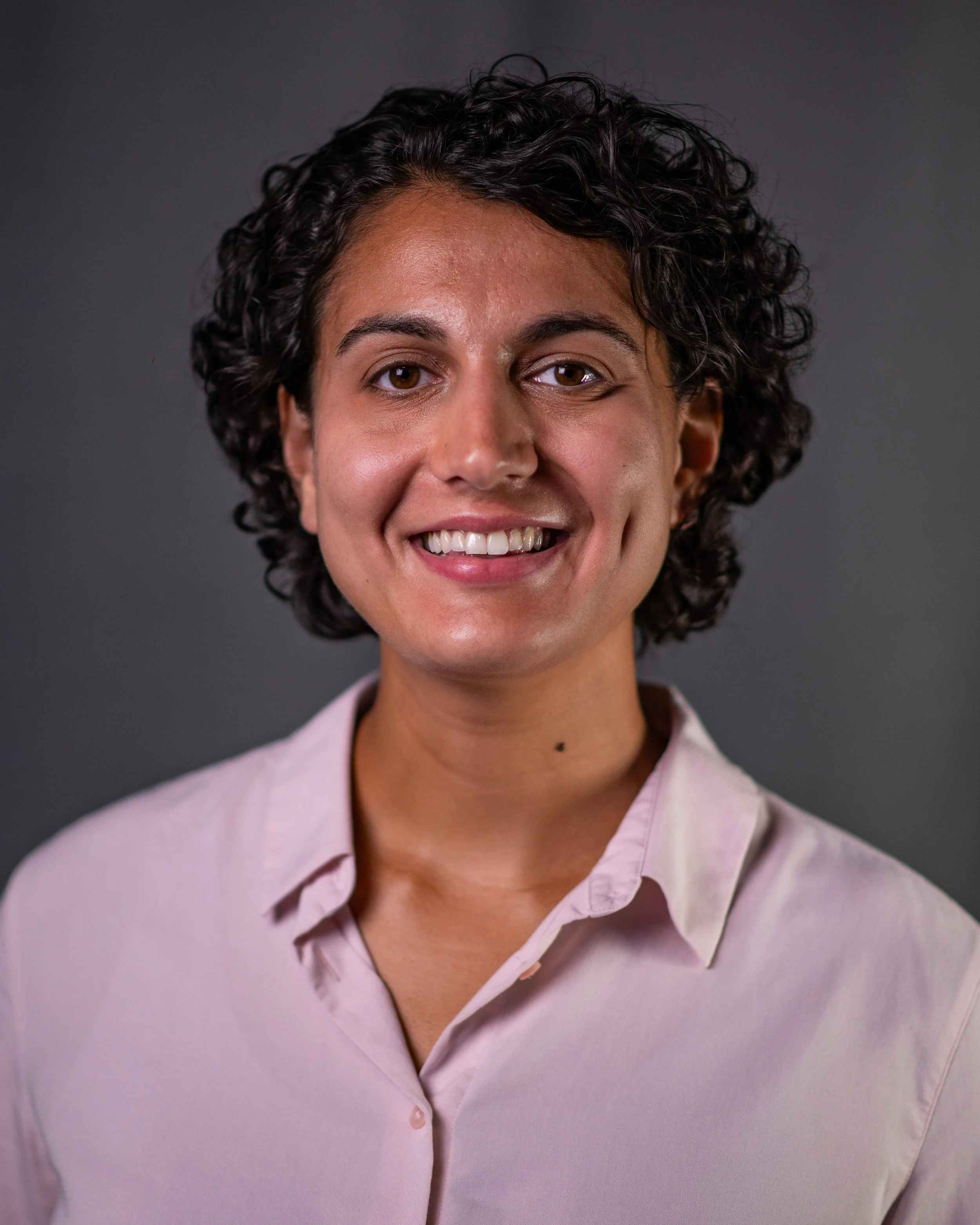 A woman with short curly dark hair, wearing a light pink collared shirt, smiling against a dark grey background.