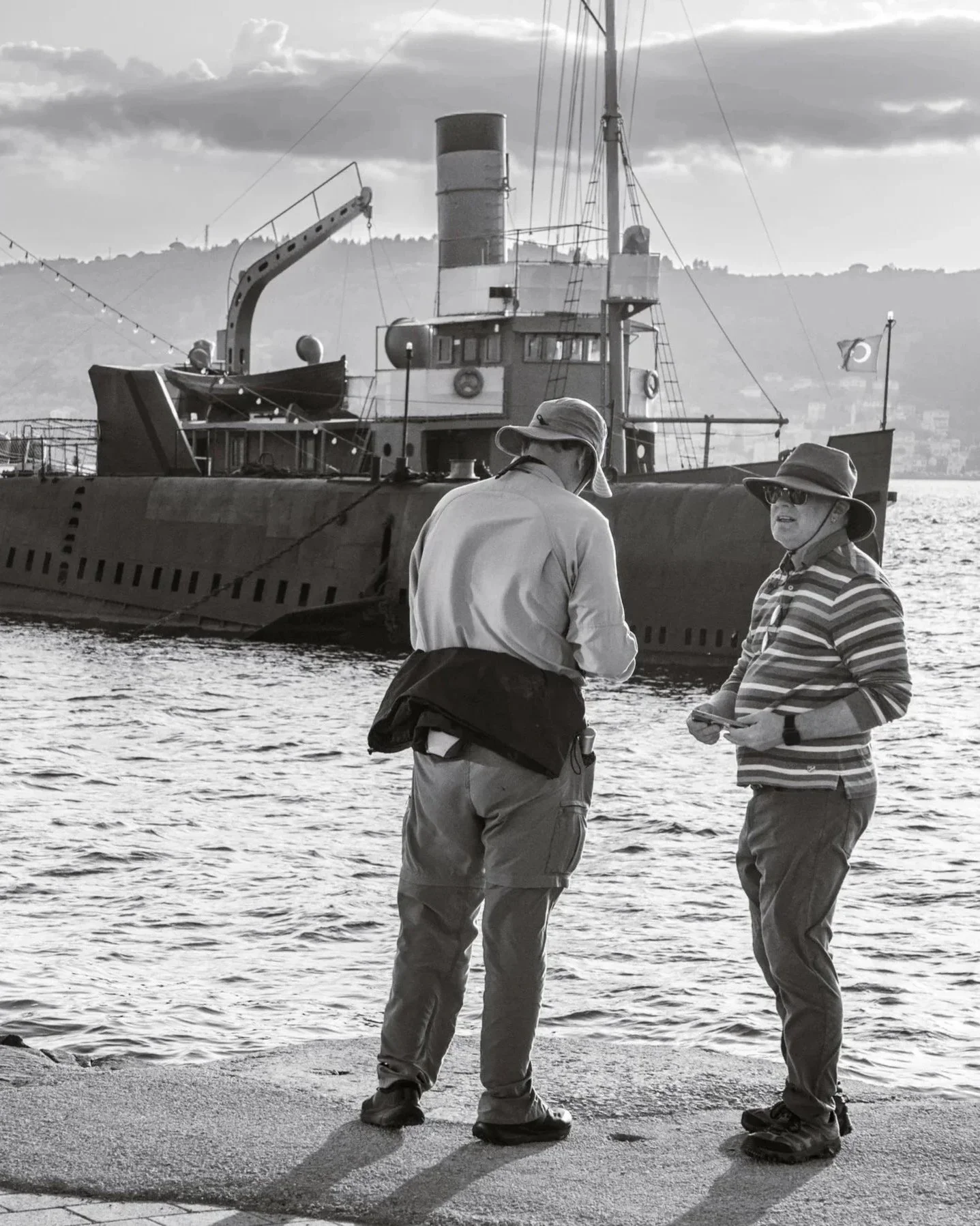 Two sun‑hatted friends linger by the water’s edge, trading quiet words as a retired submarine and ship rest behind them like relics from another century, floating in the soft, silvery light.