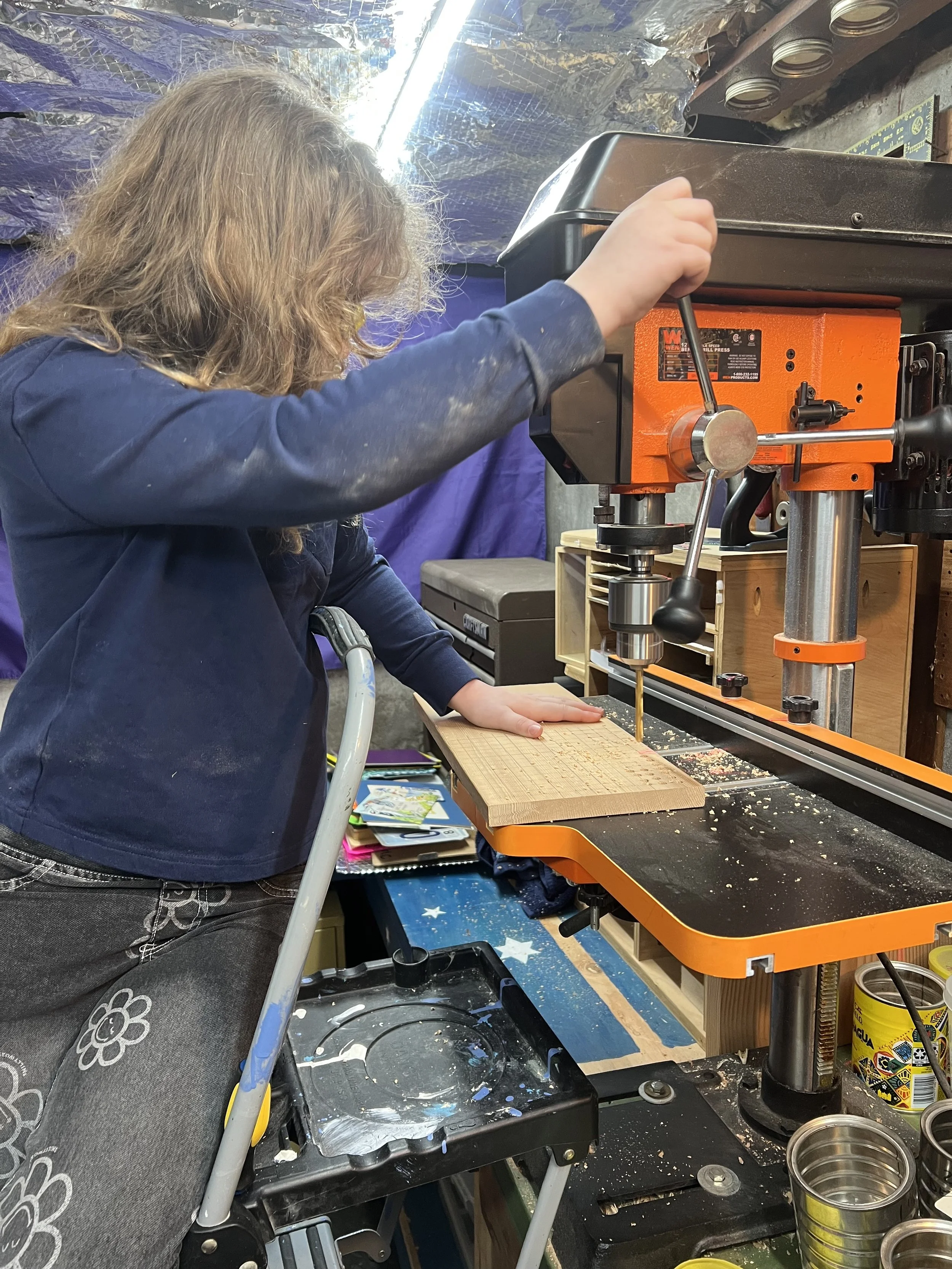 A person working with a drill press in a workshop. They are wearing a dark long-sleeve shirt, and their left hand is holding a piece of wood on the table while their right hand operates the drill. The workshop has various tools and supplies visible, including cans and papers.
