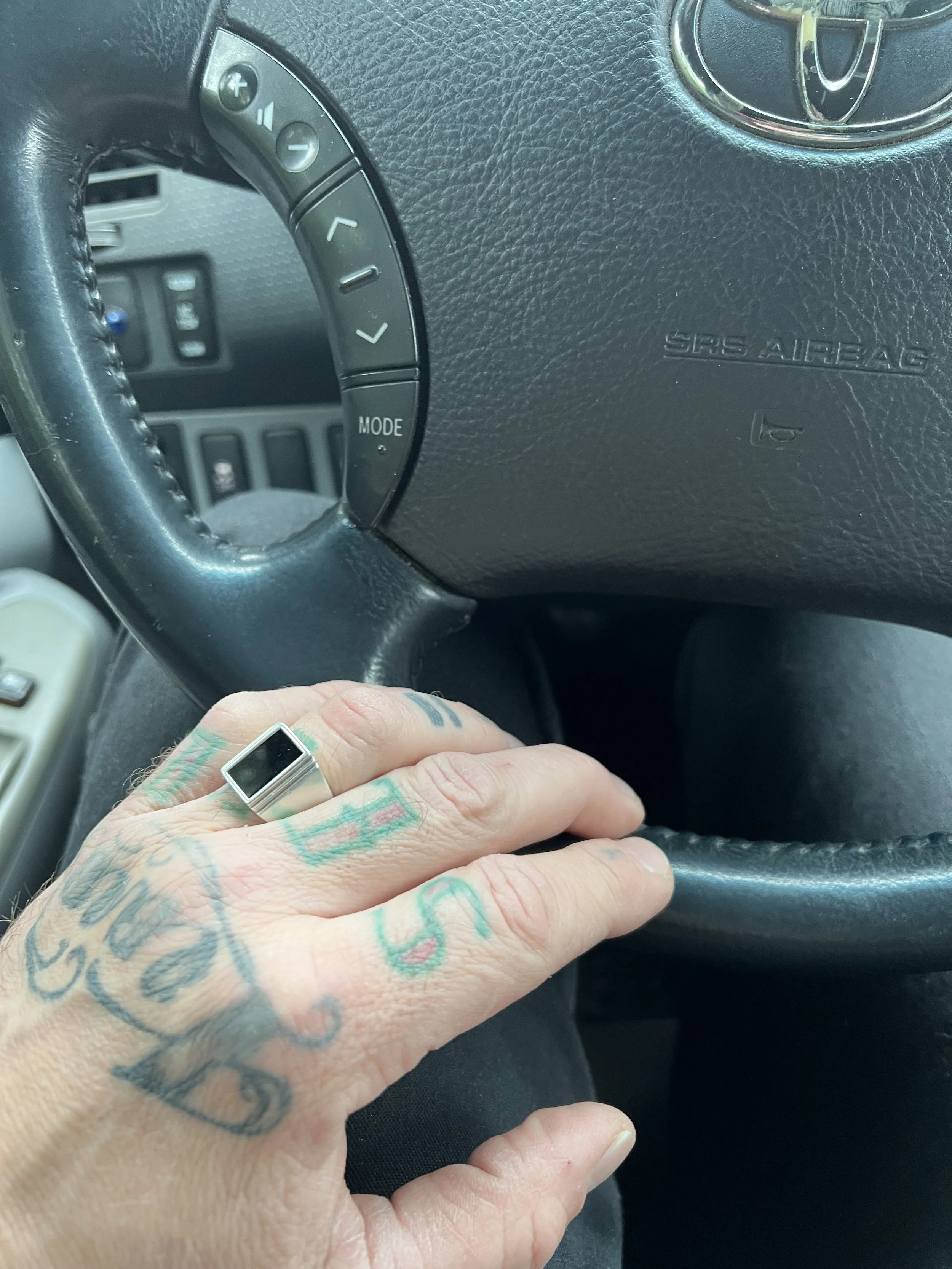 A tattooed hand resting on a black steering wheel inside a car, with part of the car's dashboard visible. The steering wheel has buttons, and the car's airbag cover is marked with 'SRS AIRBAG'.