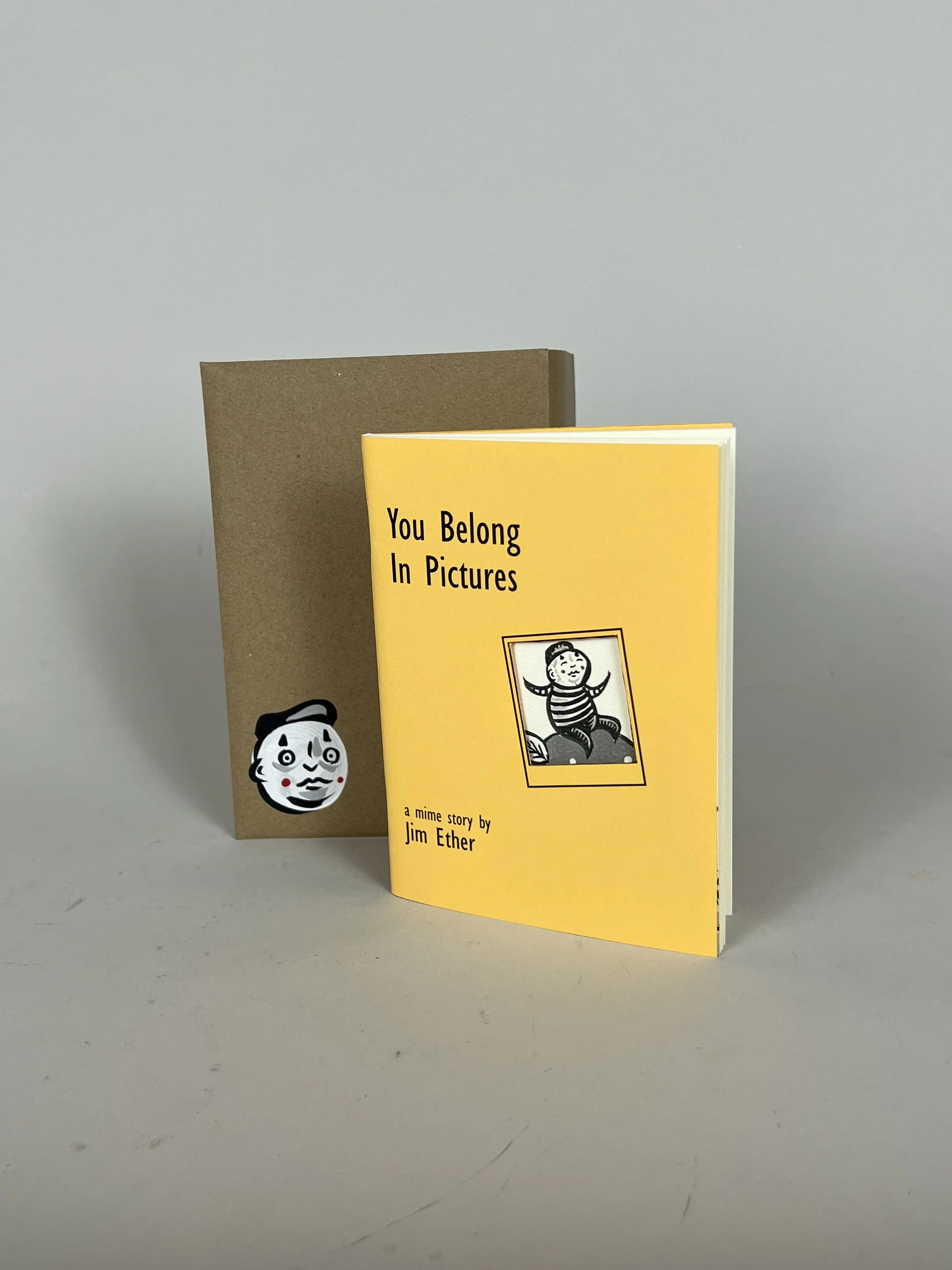 A small book titled "You Belong In Pictures" with a black and white illustration of a smiling boy on a striped shirt, placed in front of a brown paper bag featuring a sticker of a smiling cartoon face. Made by Jim Ether and Scott Cannon