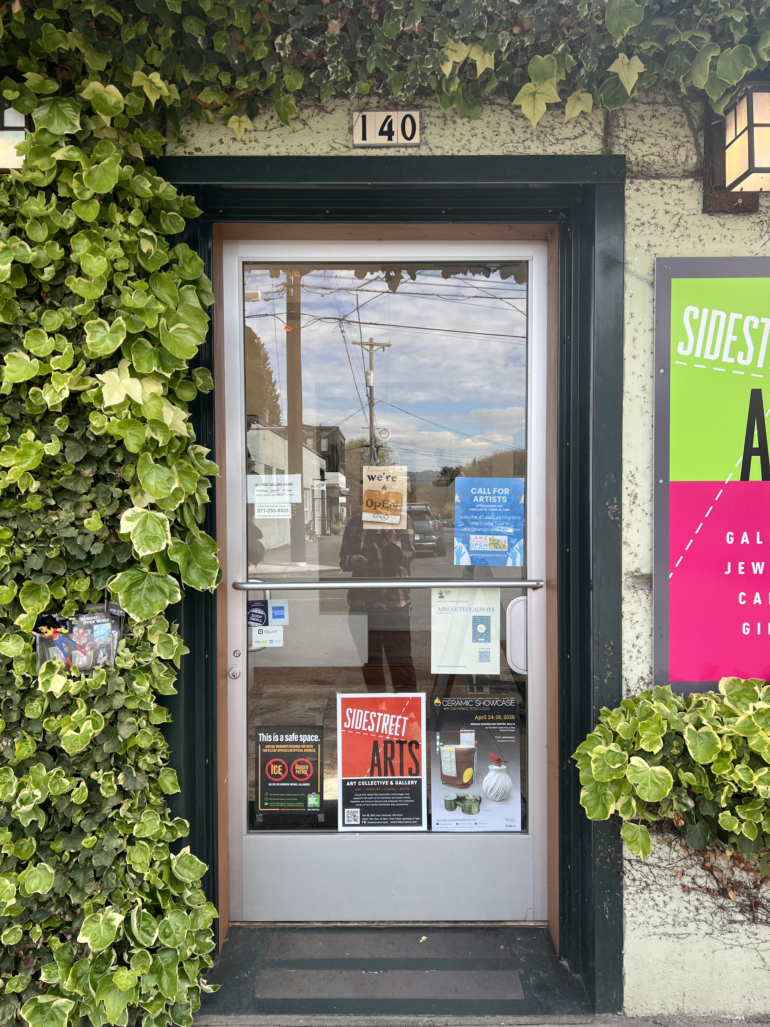 The entrance to an art gallery with the address 140 above the door. The door has posters for art events, including Sidestreet Arts. There are green vines on both sides of the entrance and a sign that says 'We're Open' in the window.