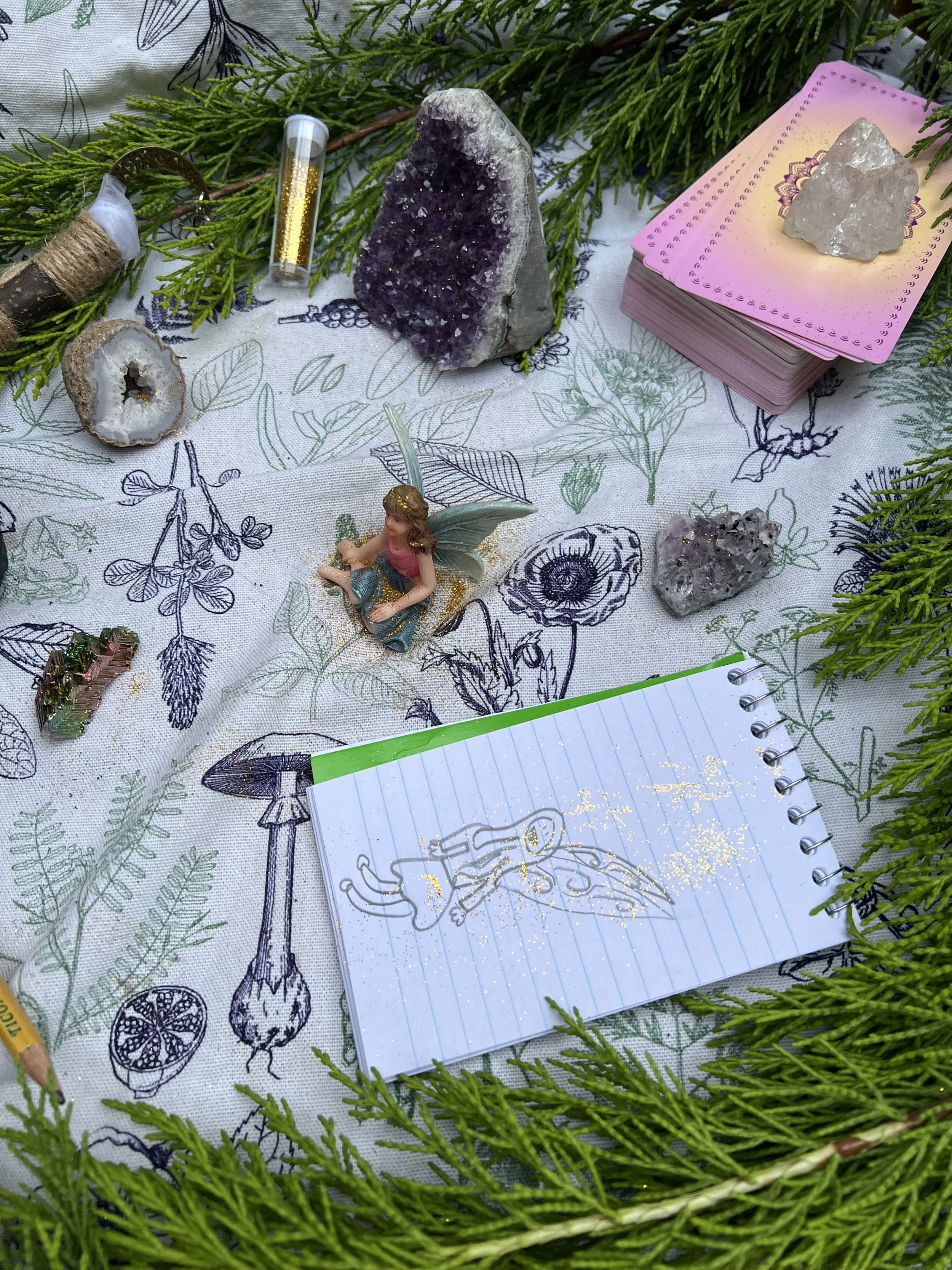 A collection of crystals, a fairy figurine, and tarot cards surrounded by greenery on a decorated tablecloth.
